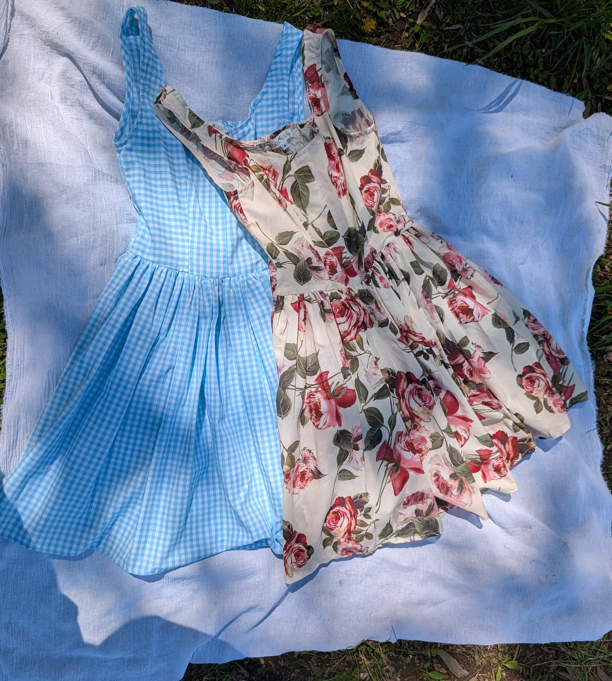 Two dresses, one blue checkered and one floral, laid out on a white sheet outdoors.