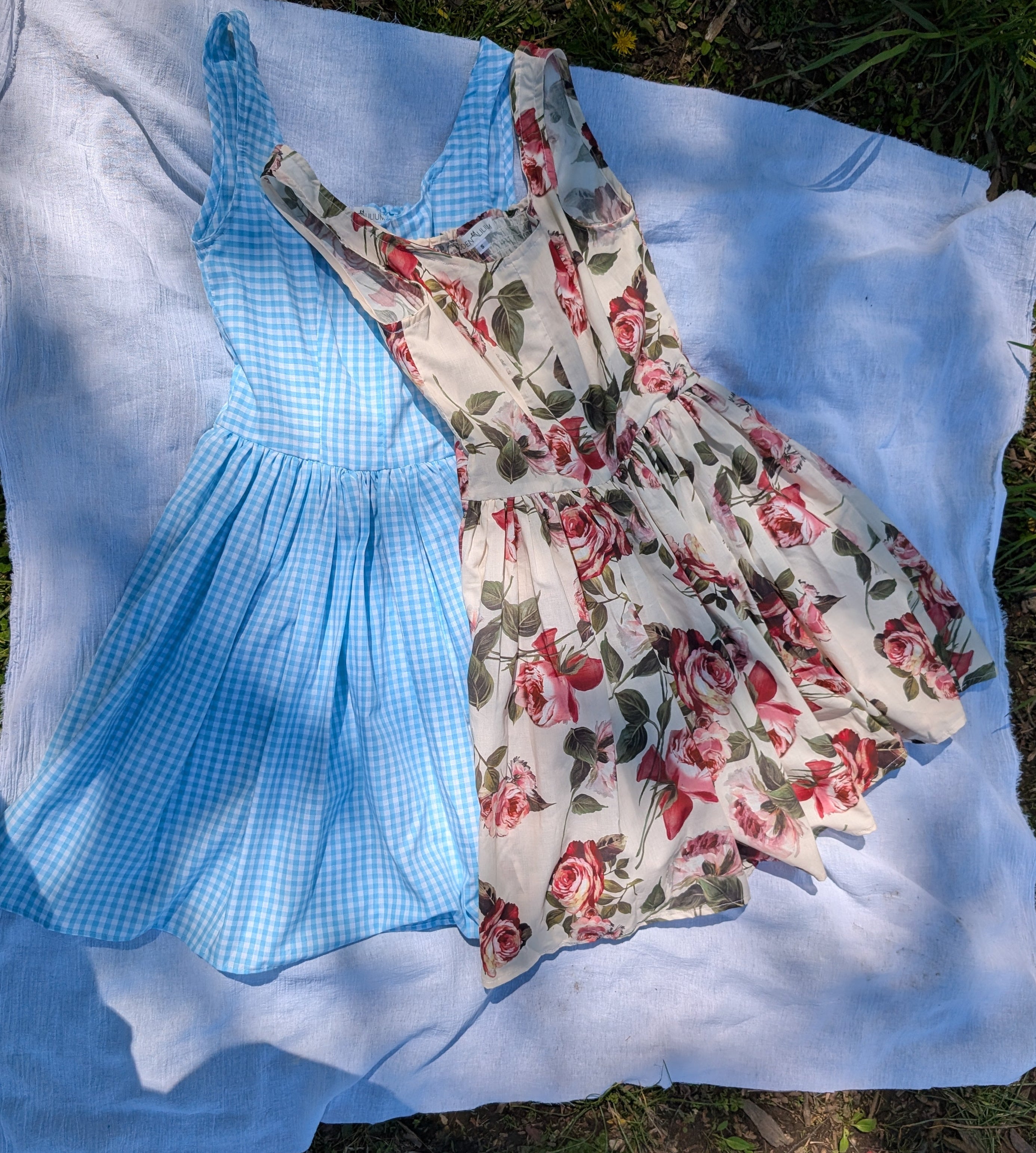 Two dresses, one blue checkered and one floral, laid out on a white sheet outdoors.