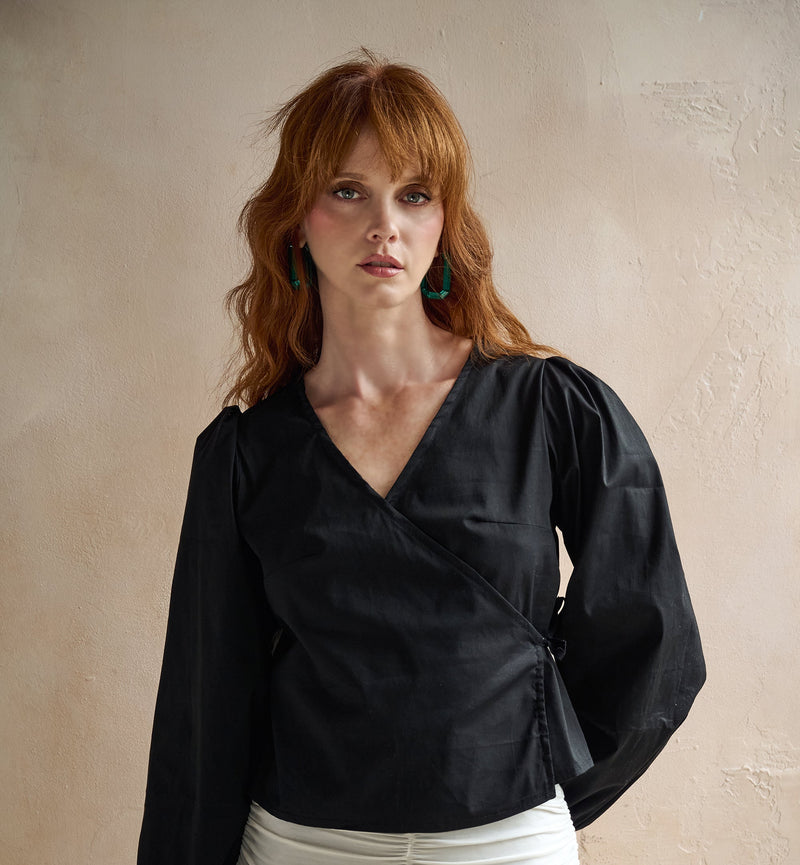 A woman with red hair wearing a black long-sleeve wrap-style blouse with puffed shoulders and a white ruched skirt with floral embroidery, standing against a textured beige wall.