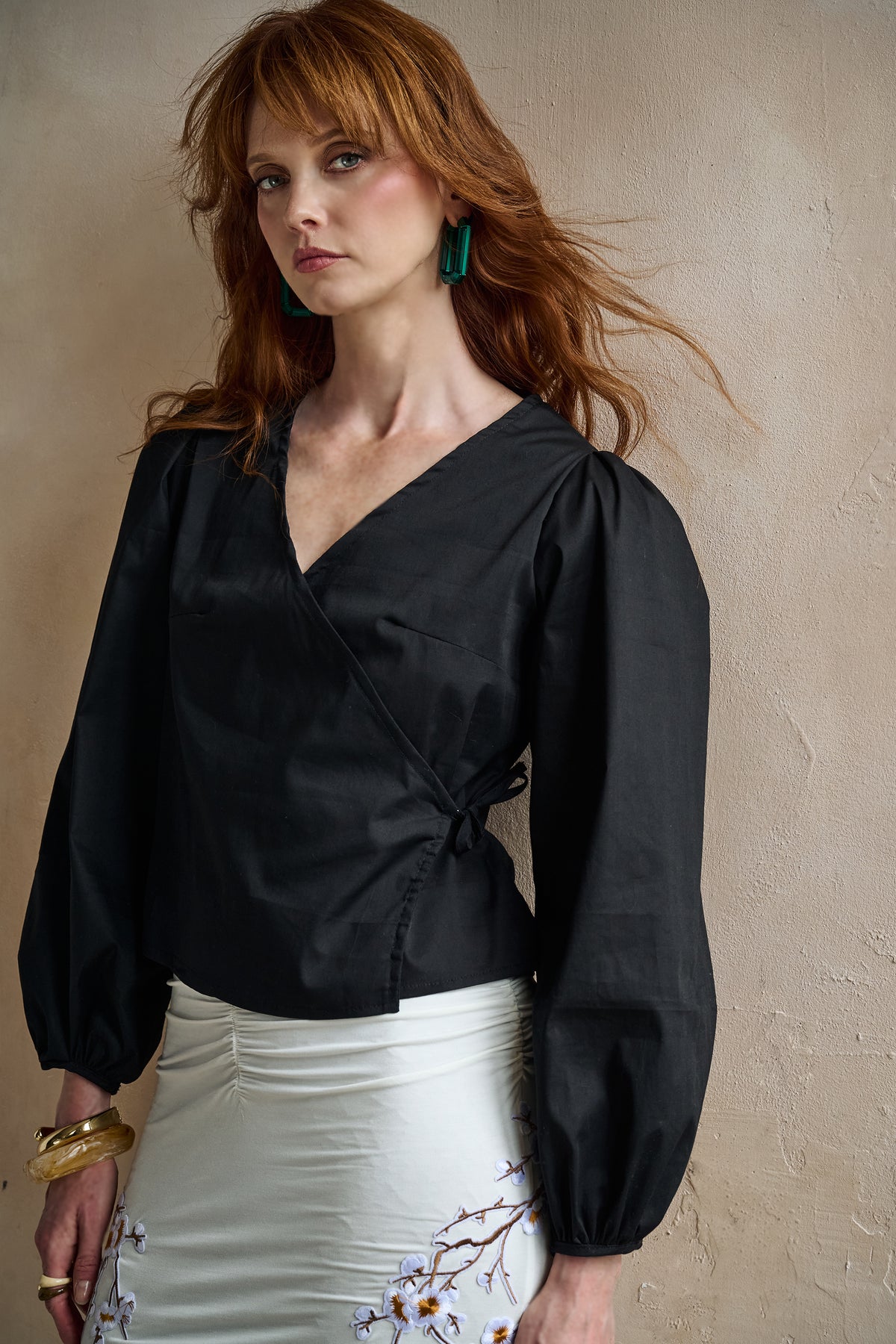 A woman with red hair wearing a black long-sleeve wrap-style blouse with puffed sleeves and a white skirt, standing against a textured beige wall.