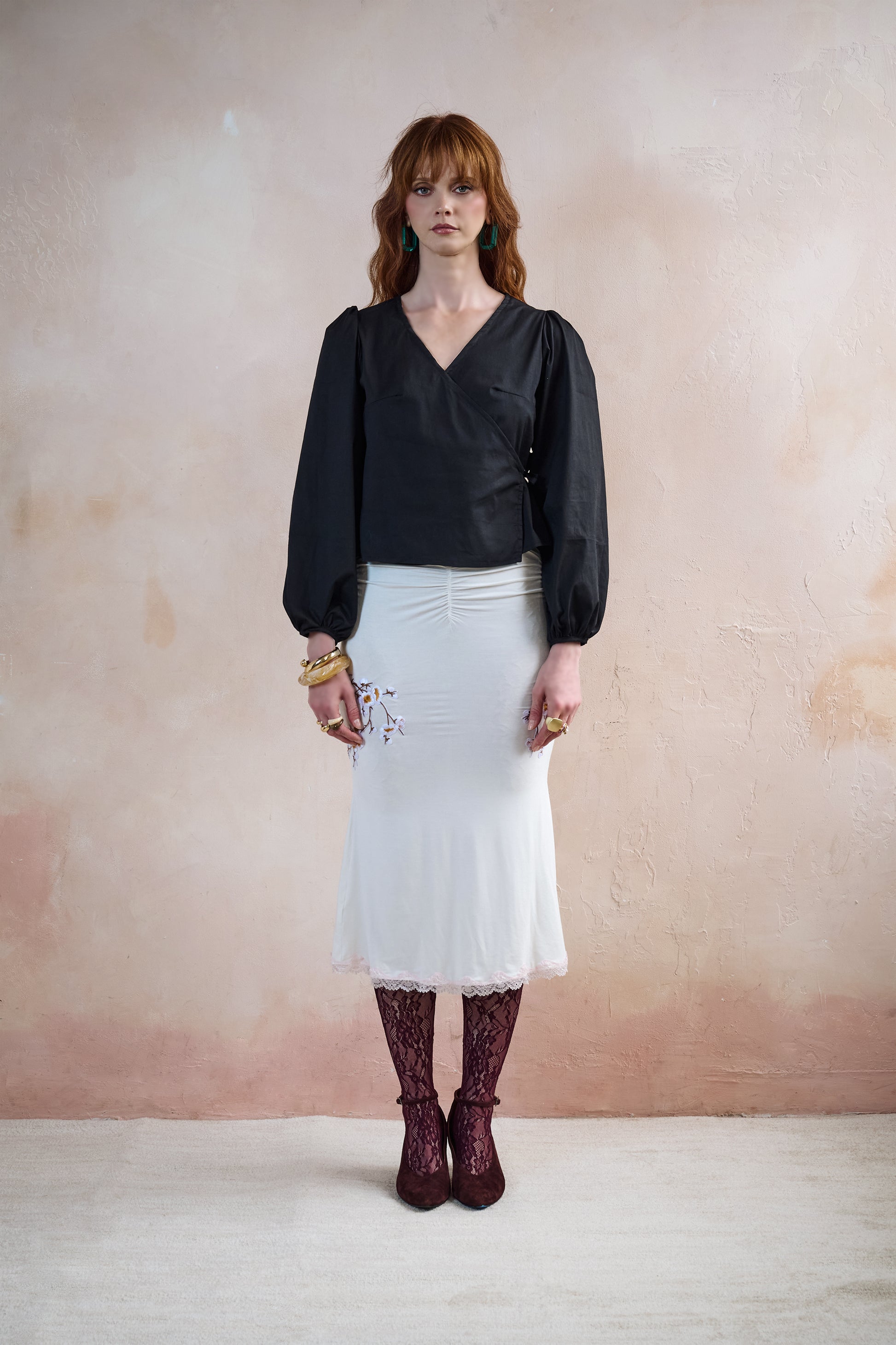 A woman stands against a textured beige wall wearing a black long-sleeve blouse with puffed sleeves, a white gathered midi skirt, burgundy lace-up tights, and dark burgundy ankle heel.