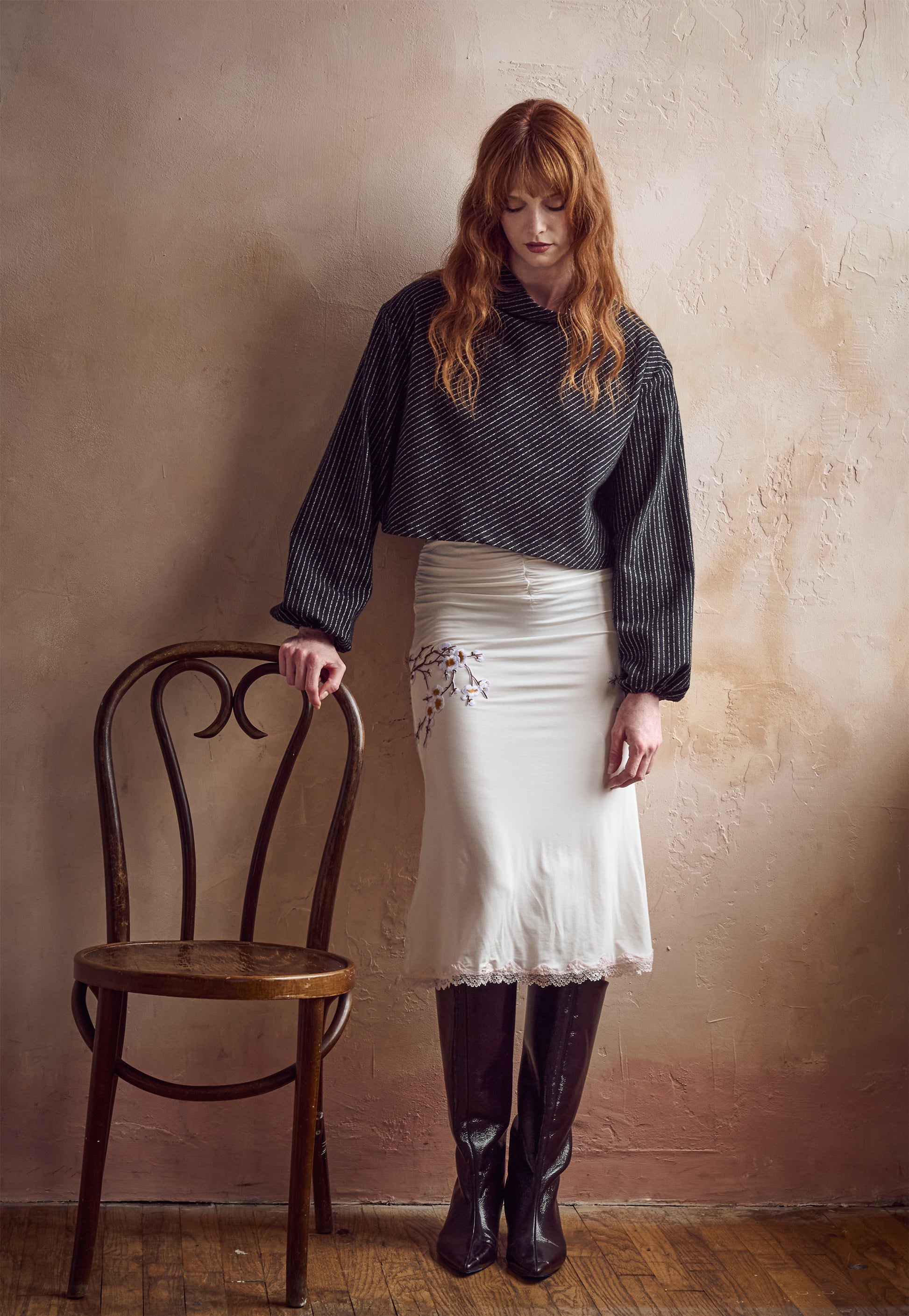 A woman with long red hair stands in a studio setting next to a wooden chair, wearing a black and white pinstripe cropped shirt, a cream skirt with floral embroidery, and dark knee-high boots against a textured beige wall.