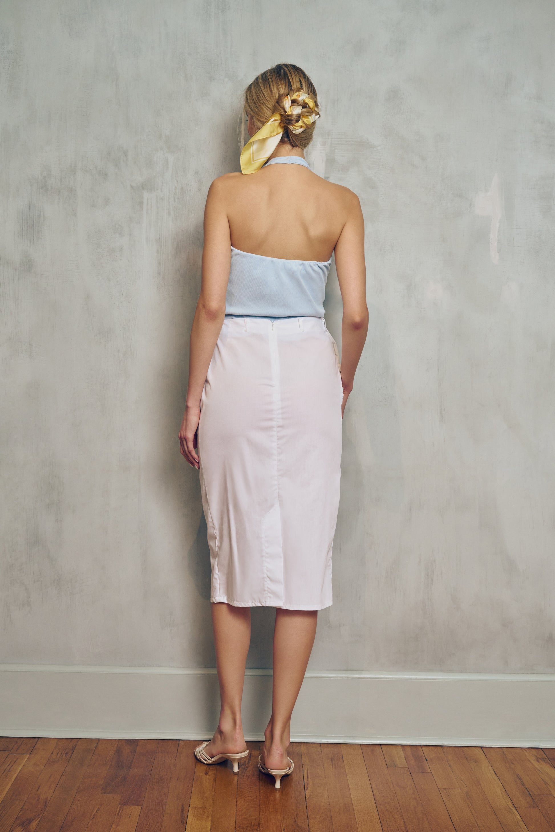 A woman wearing a halter light blue top and light pink skirt against a gray wall.