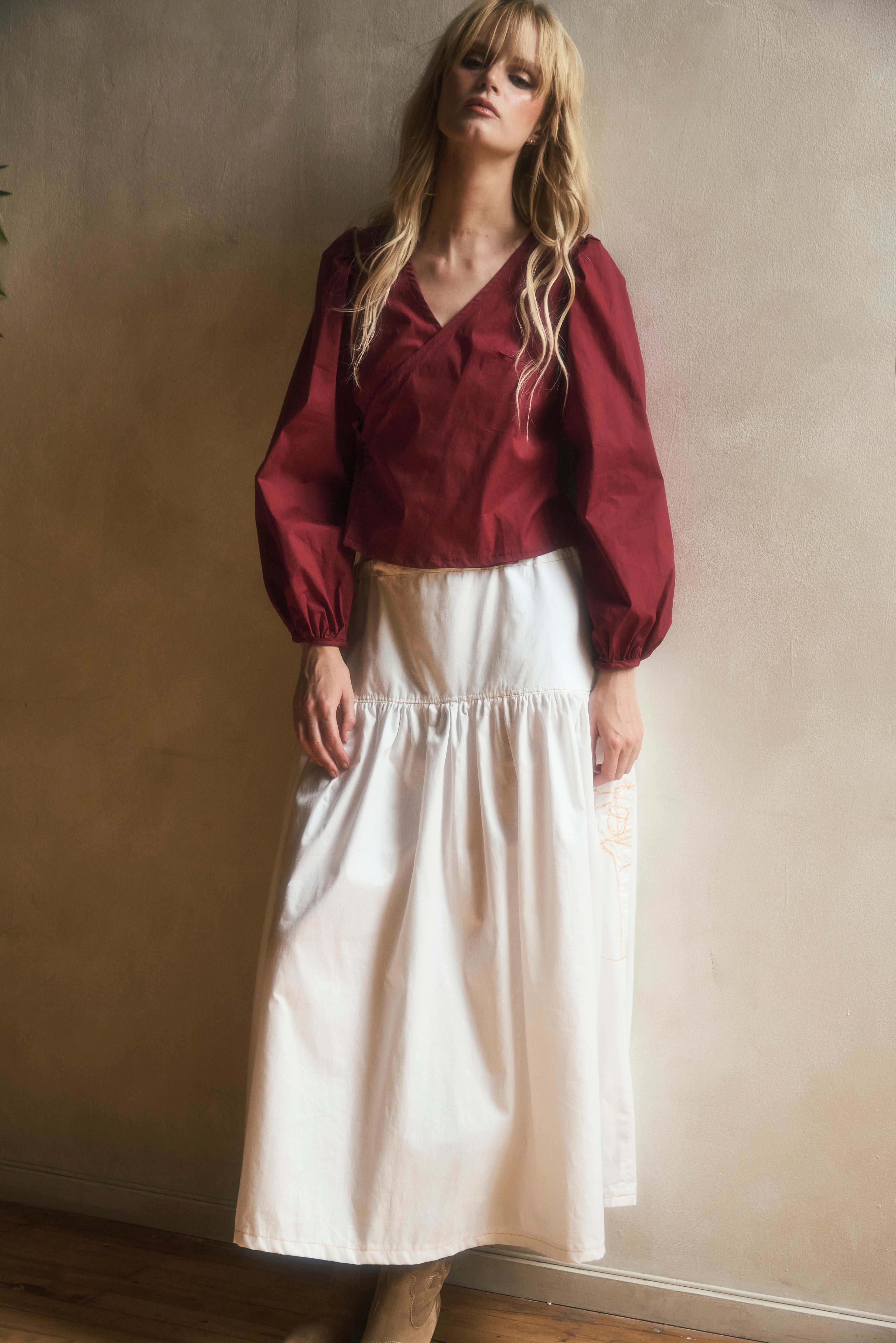 Woman wearing a burgundy blouse and white skirt against a plain wall.