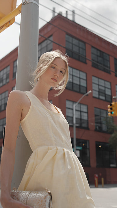 A woman wearing a sleeveless light yellow colored dress leans against a gray pole on a city street, holding a patterned clutch bag. A red brick building is visible in the background.