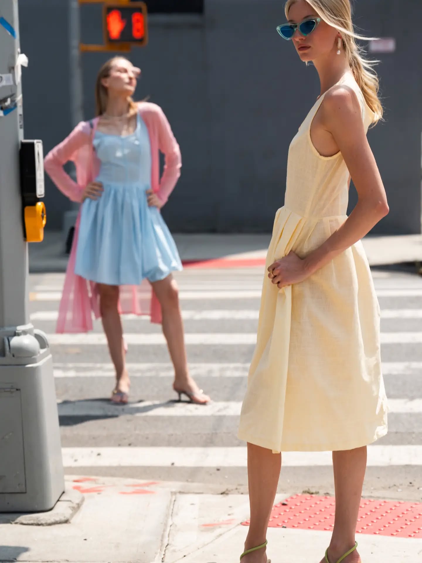 A woman in a pale yellow sleeveless midi dress and blue sunglasses stands on a city crosswalk, with another woman in a light blue dress and pink cardigan blurred in the background.