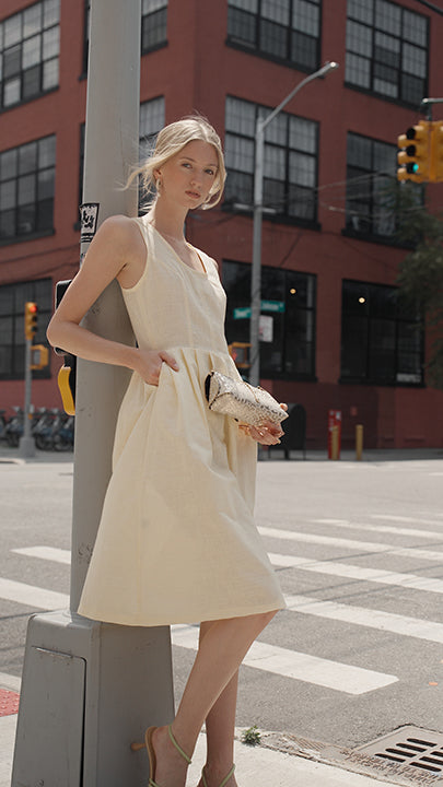 A woman stands on a city street corner wearing a sleeveless buttercup yellow colored midi dress with a gathered waist and side pocket, holding a metallic clutch. The background features a red brick building and a gray utility pole.