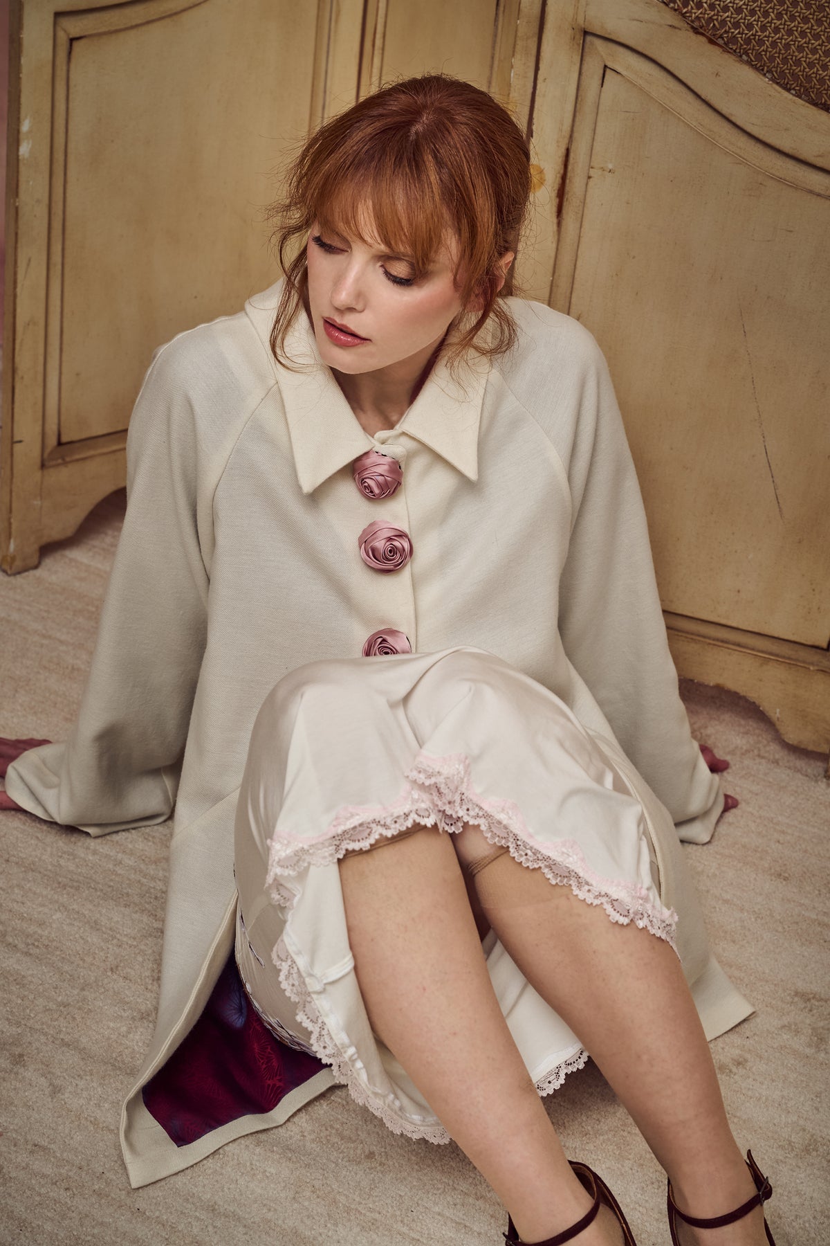 A woman with red hair sits on a light-colored floor in front of a wooden cabinet, wearing a cream-colored coat with pink fabric roses on the collar and hem, and a lace-trimmed skirt.