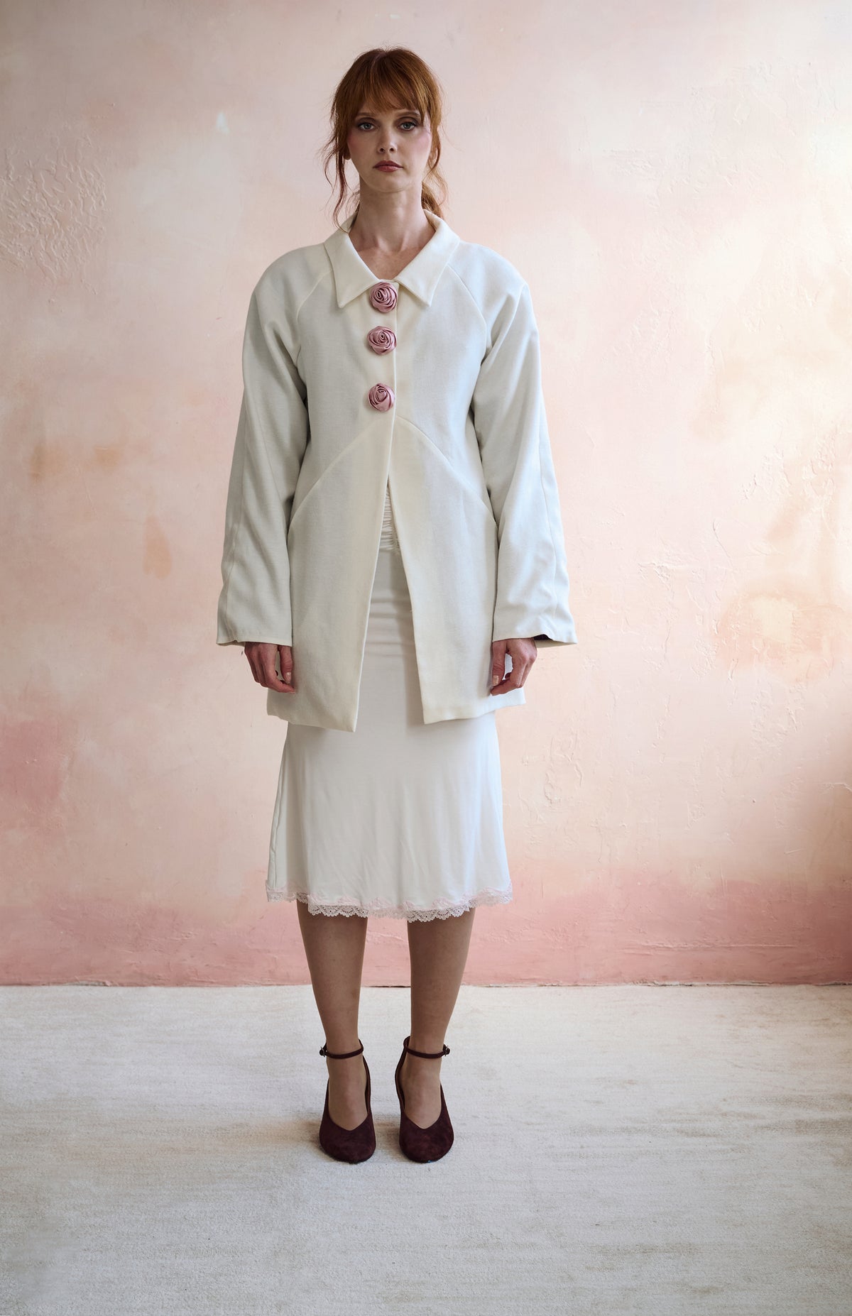 A woman stands against a soft pink textured wall wearing a cream-colored jacket with pink decorative buttons and a matching cream midi skirt, paired with dark brown heels.