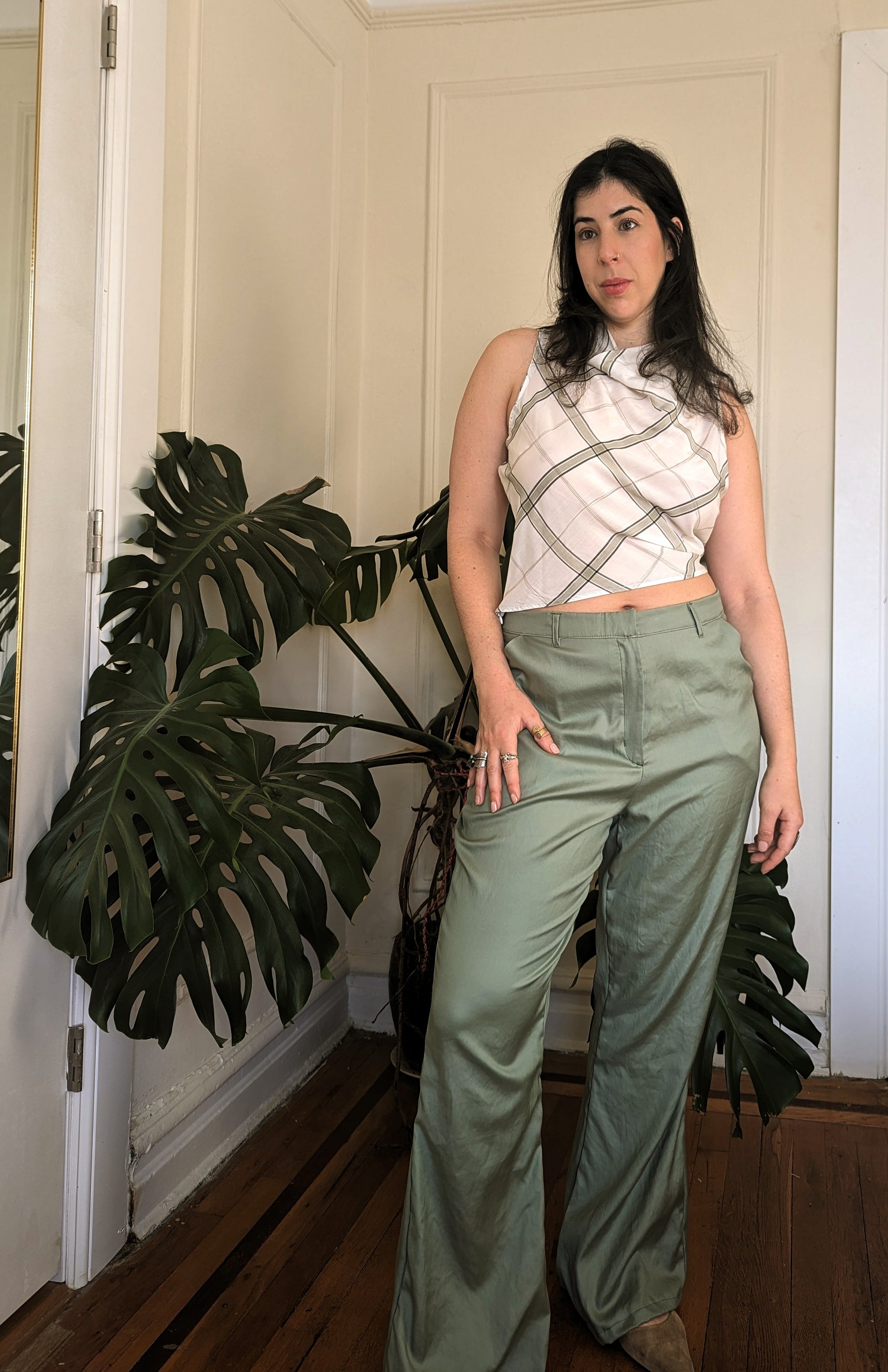 Woman wearing a sleeveless plaid top and high-waisted pants standing indoors with a plant in the background.