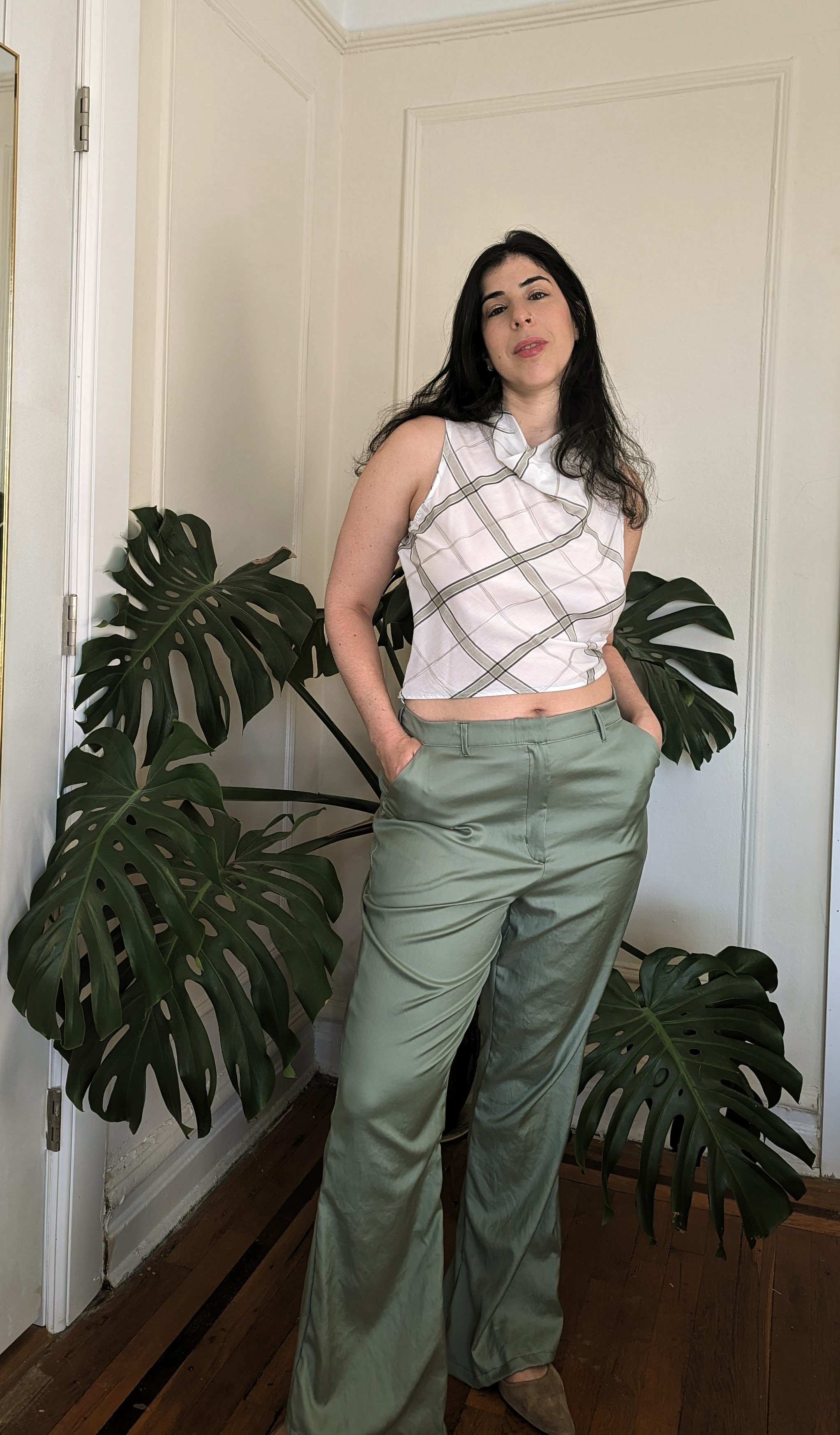 Woman standing indoors wearing plaid sleeveless top and green pants with a plant in the background