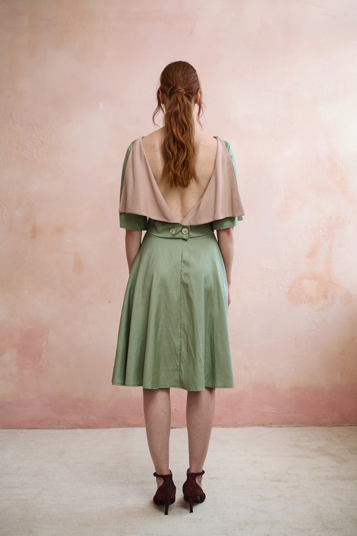 A woman stands with her back to the camera wearing a sage green midi skirt with a structured waist and a open back style top, set against a textured pinkish-beige wall.
