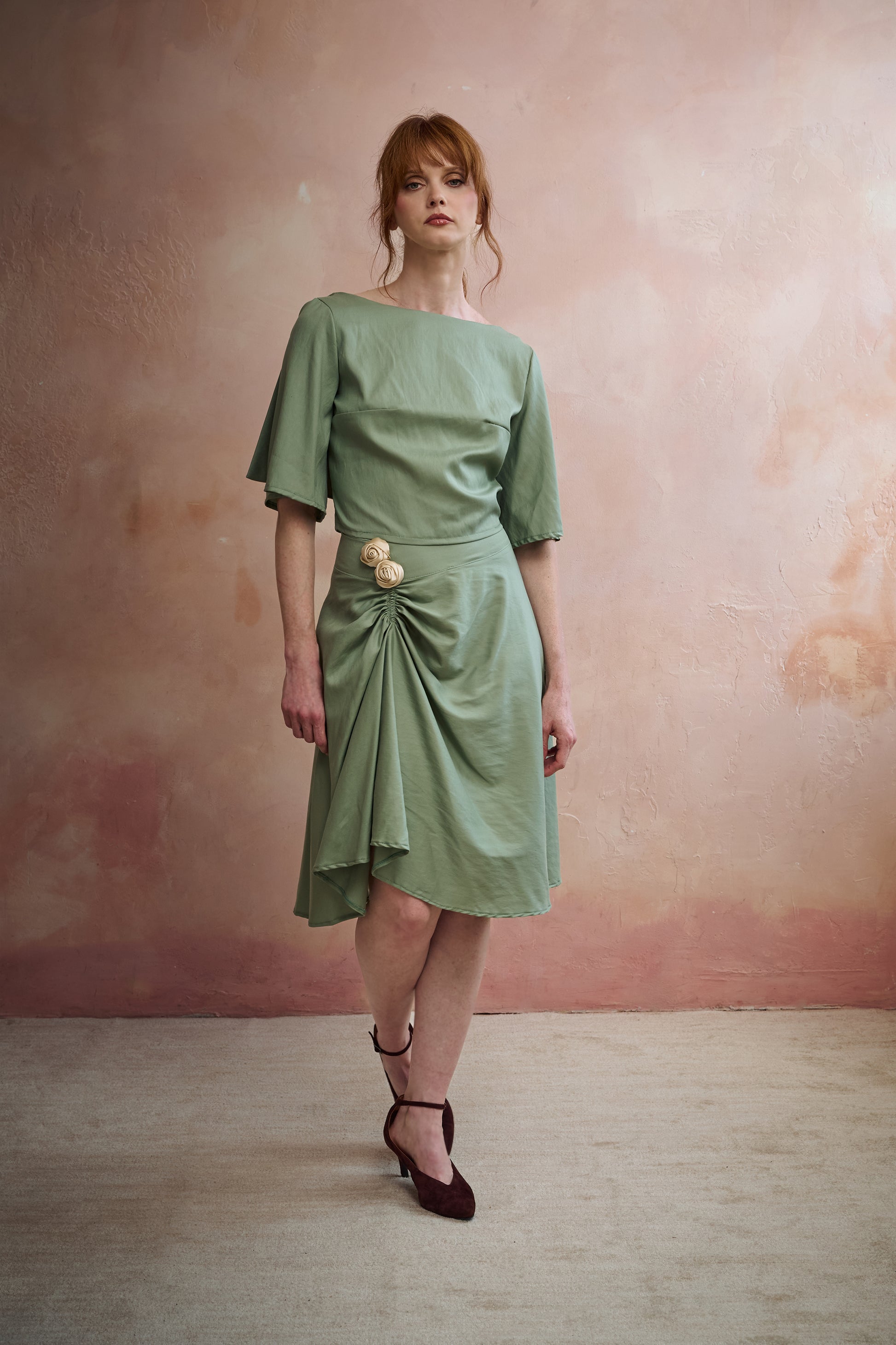 A woman models a sage green midi skirt with a draped, gathered detail and a decorative beige flower accent at the waist, set against a textured pinkish-beige studio wall.