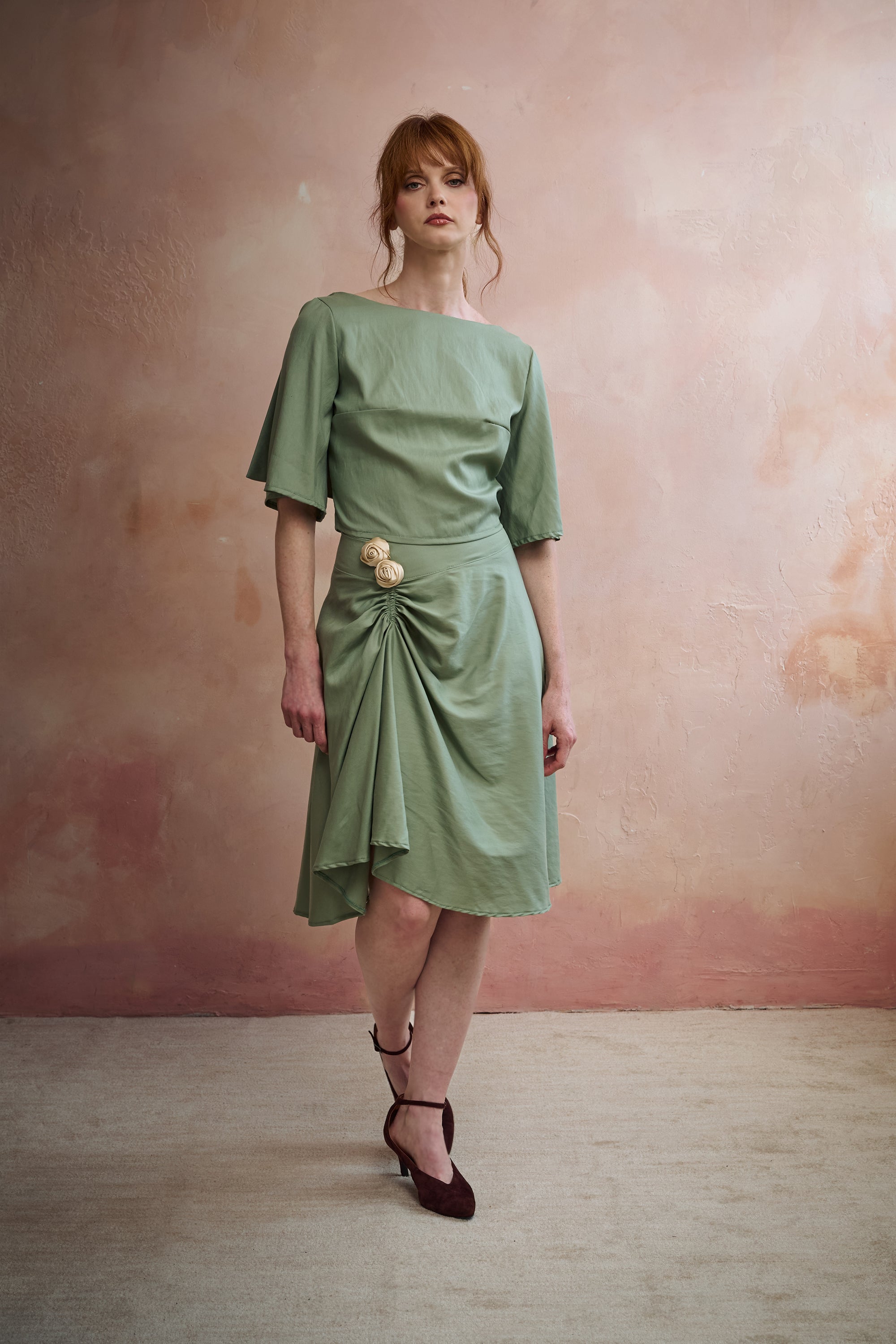 A woman models a sage green midi skirt with a draped, gathered detail and a decorative beige flower accent at the waist, set against a textured pinkish-beige studio wall.