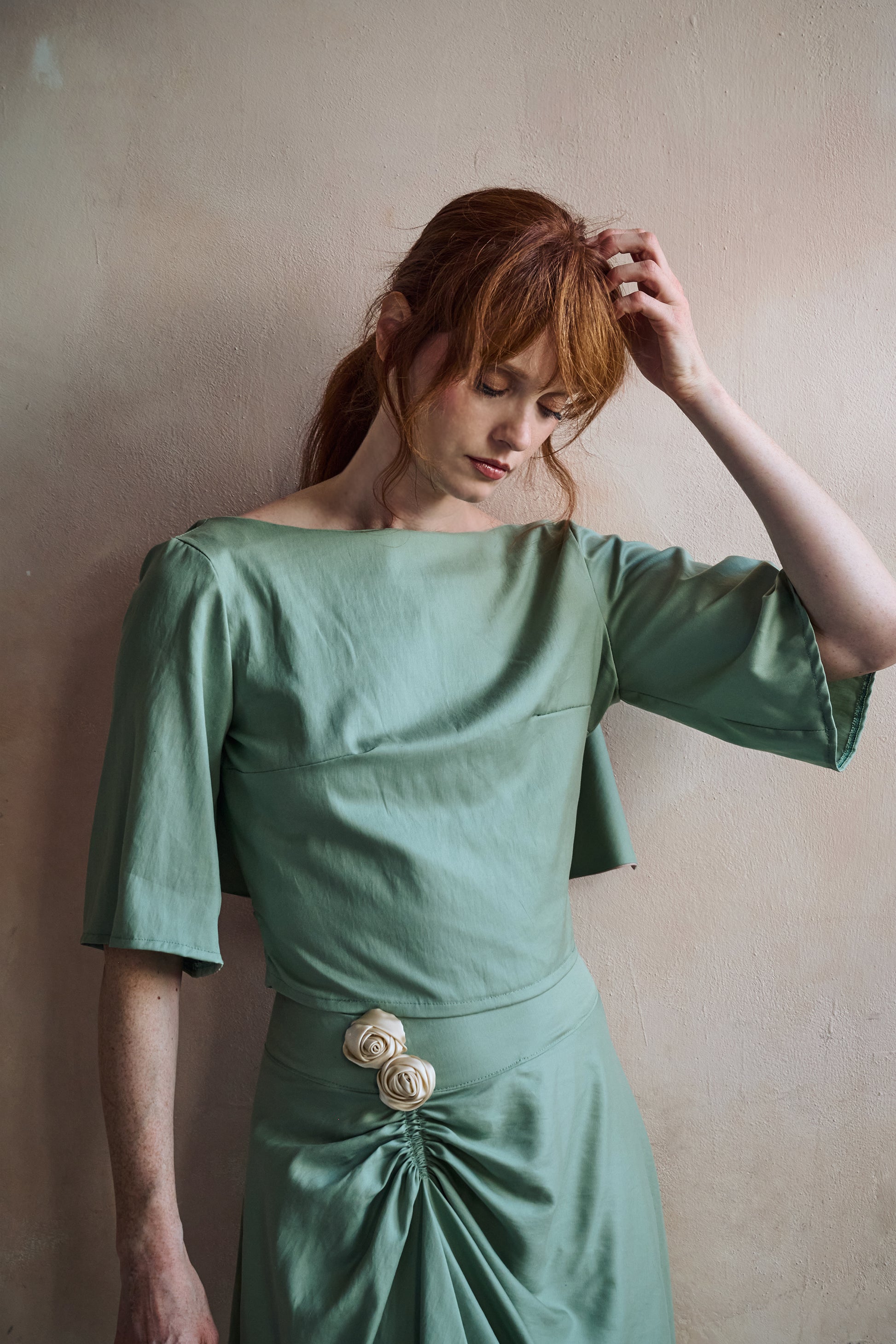 A woman with red hair wearing a sage green shirt with elbow-length sleeves and a skirt with gathered waist detail, posing against a textured beige wall.