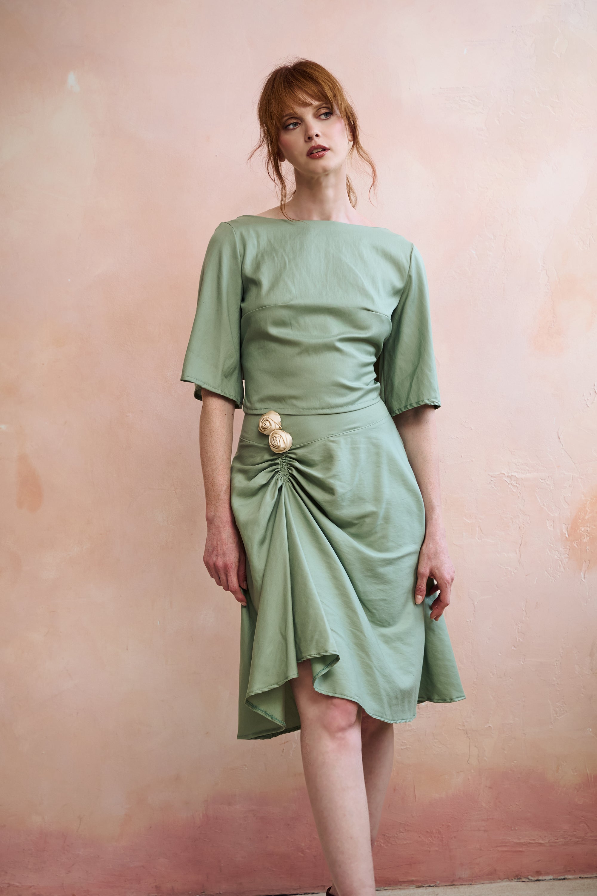 A woman modeling a sage green midi skirt with short sleeves top, a boat neckline, and a gathered skirt detail accented with three large cream rosette, posed against a textured pinkish-beige wall