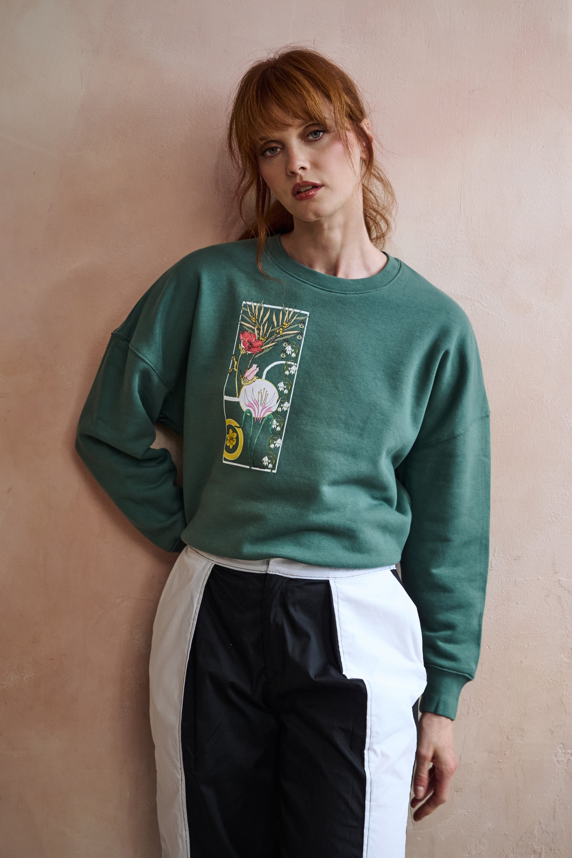 A woman with red hair wearing a green crewneck sweatshirt with a colorful floral graphic on the front, paired with white and black color-blocked pants, posing against a textured beige wall.