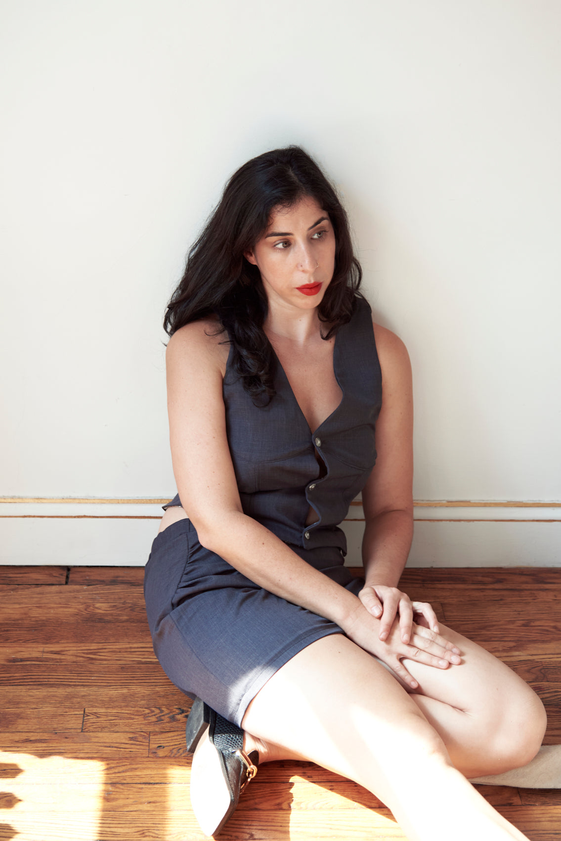 Woman in a gray vest and skirt sitting on a wooden floor with a white wall background