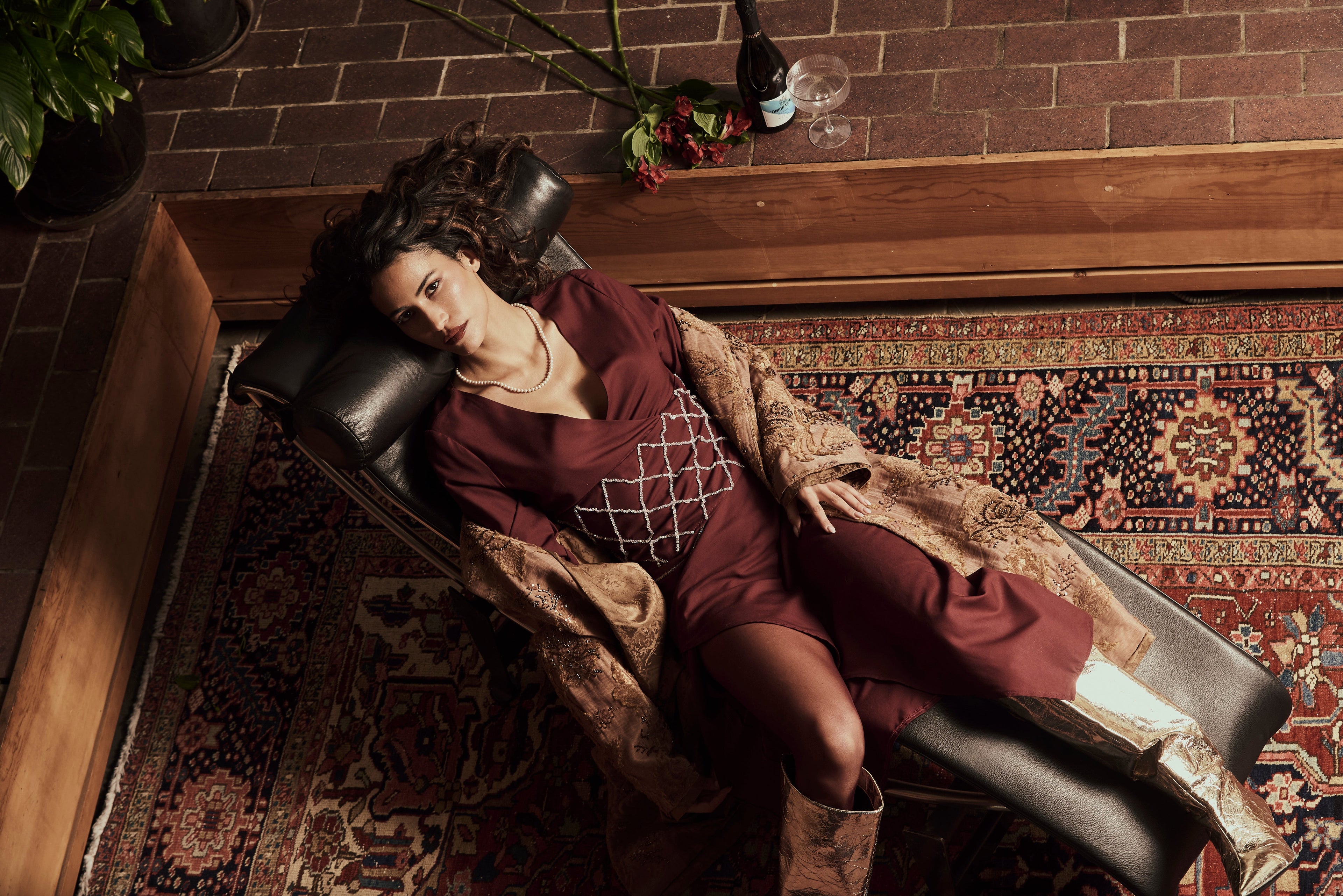 A woman in a burgundy wine-embroidered top and a skirt set, and coat lying on a leather chair in a room with a patterned rug.