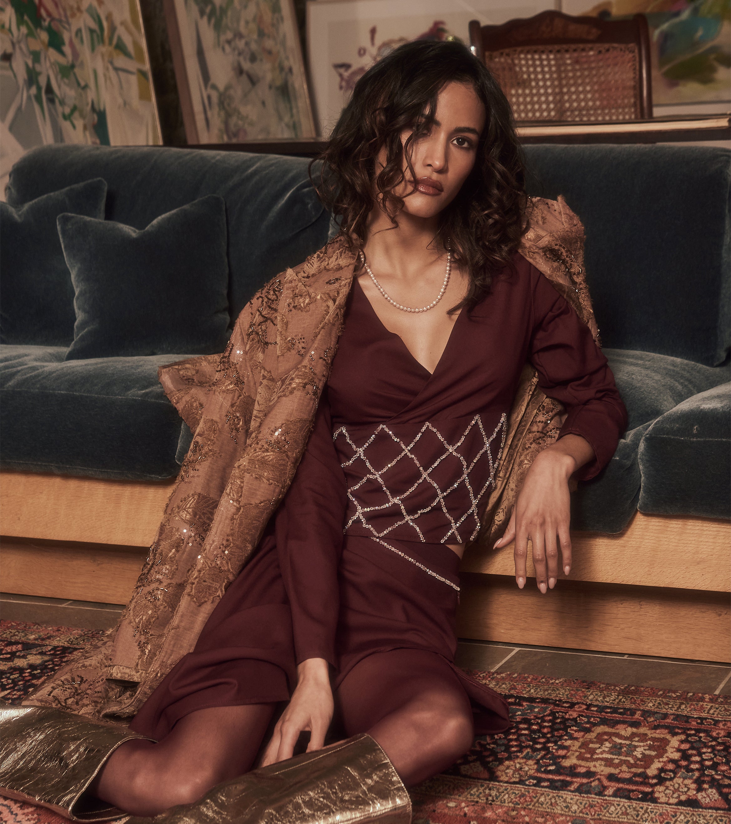 Woman in a burgundy set of long-sleeved shirt and skirt and patterned coat sitting on a blue couch.