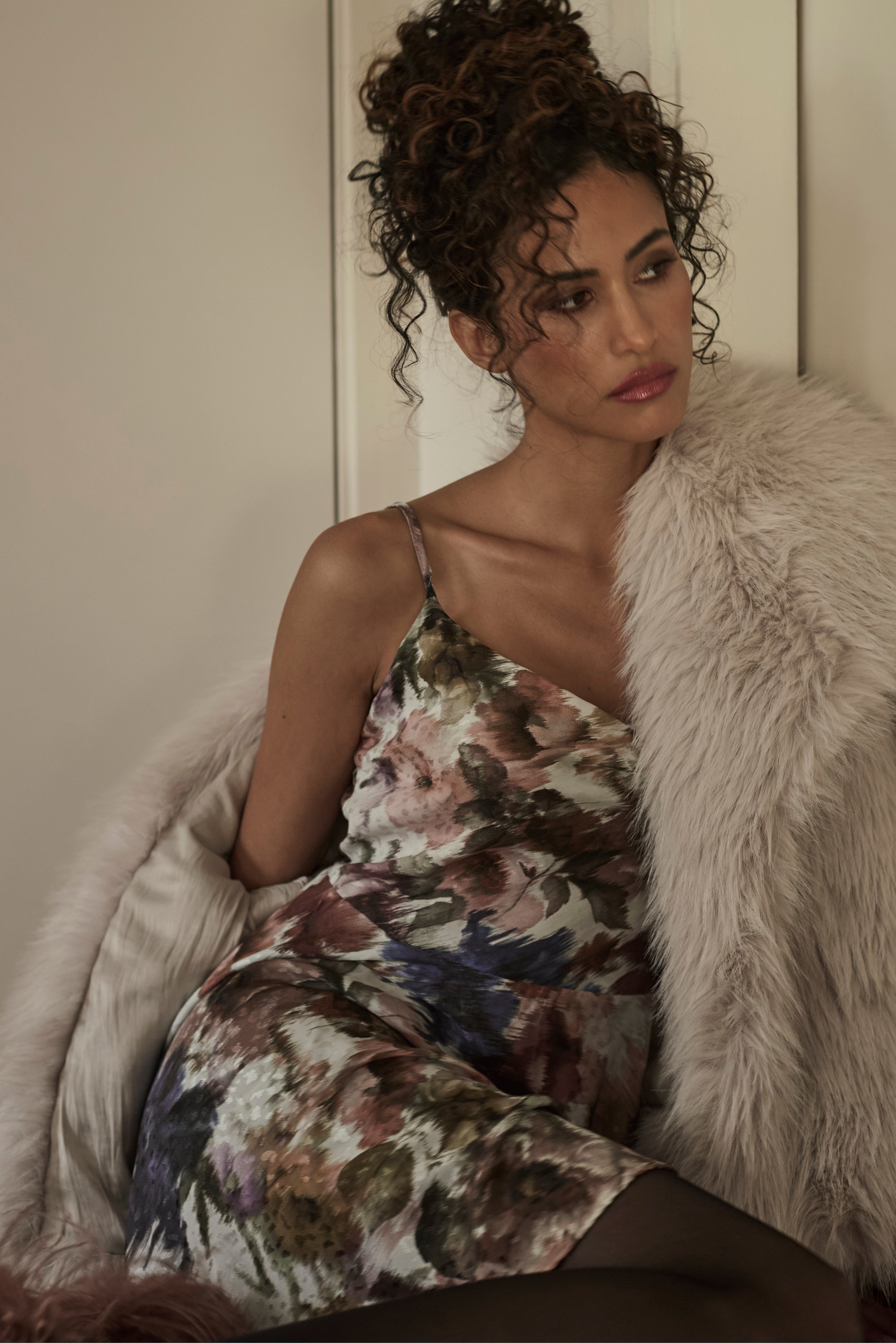 Woman in a floral dress sitting on a faux fur coat with a neutral background