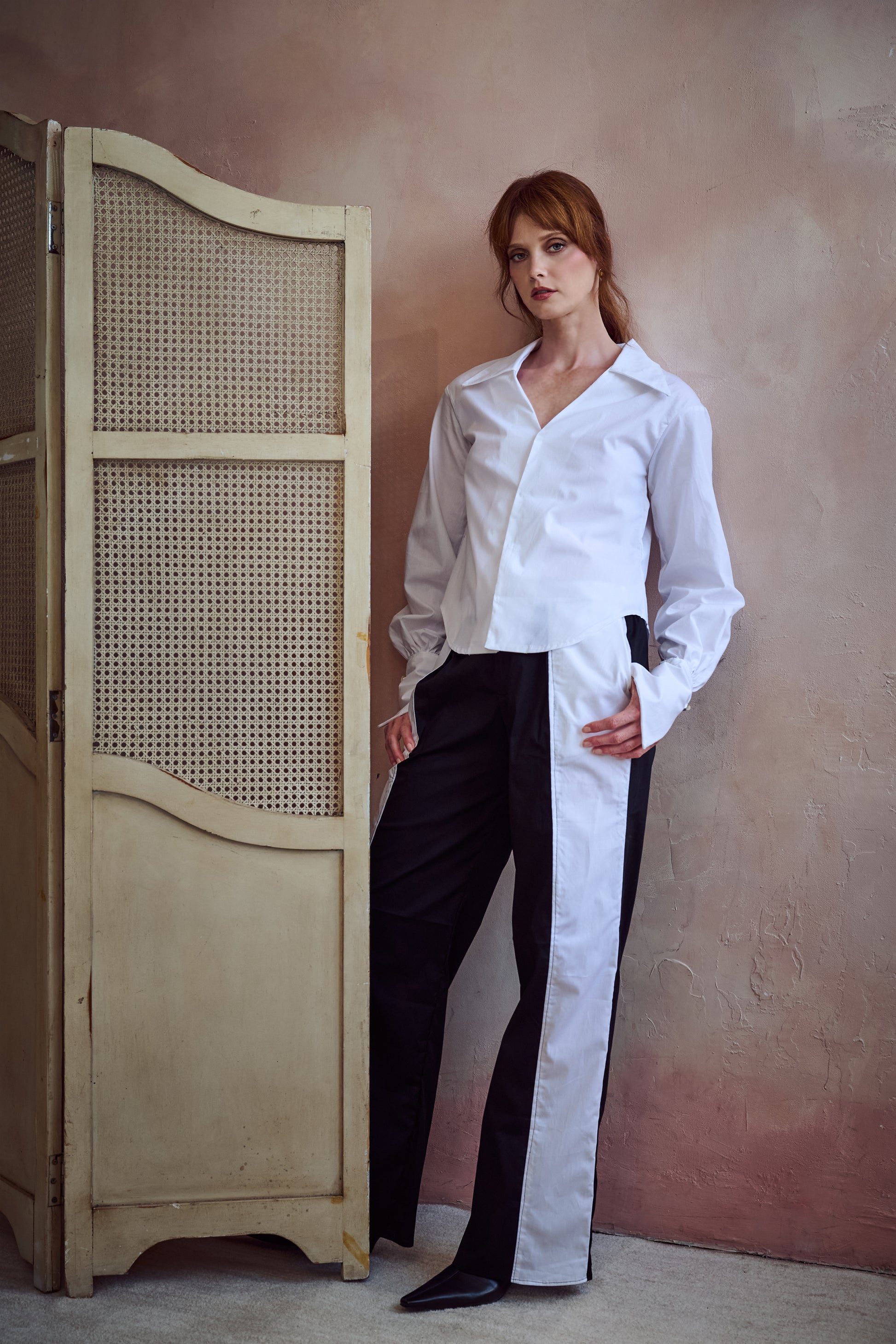 A woman stands indoors wearing a white long-sleeve button-up shirt and black-and-white wide-leg trousers, posed beside a cream folding screen against a textured warm beige wall.