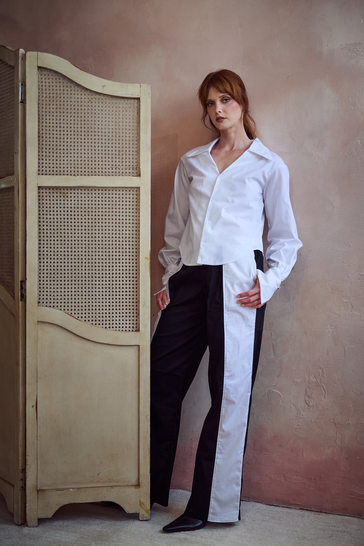 A woman stands indoors wearing a white long-sleeve button-up shirt and black-and-white wide-leg trousers, posed beside a cream folding screen against a textured warm beige wall.