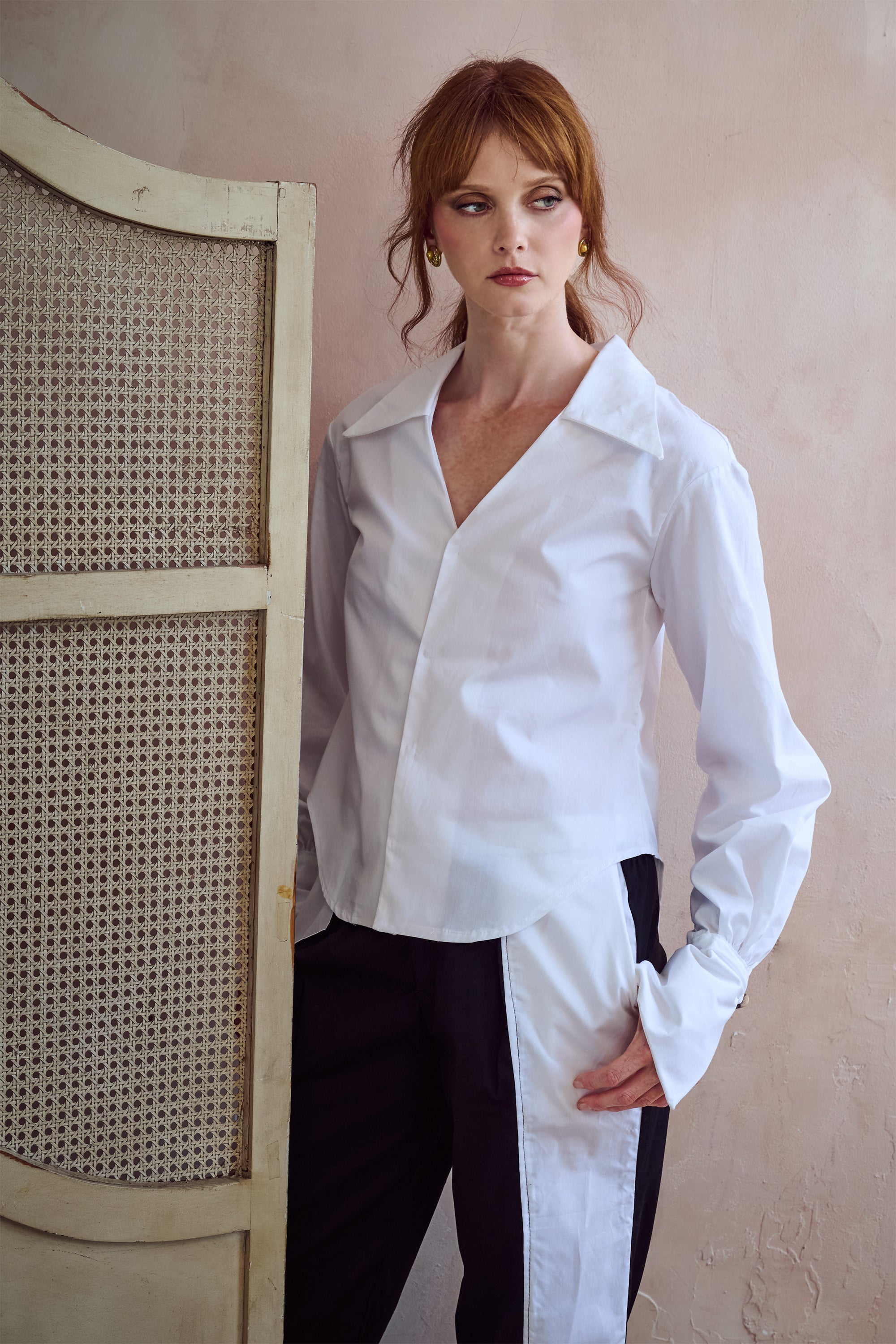 A woman with red hair models a white long-sleeve button-down shirt with black side panels, paired with black pants, standing next to a light-colored cane screen against a textured beige wall.