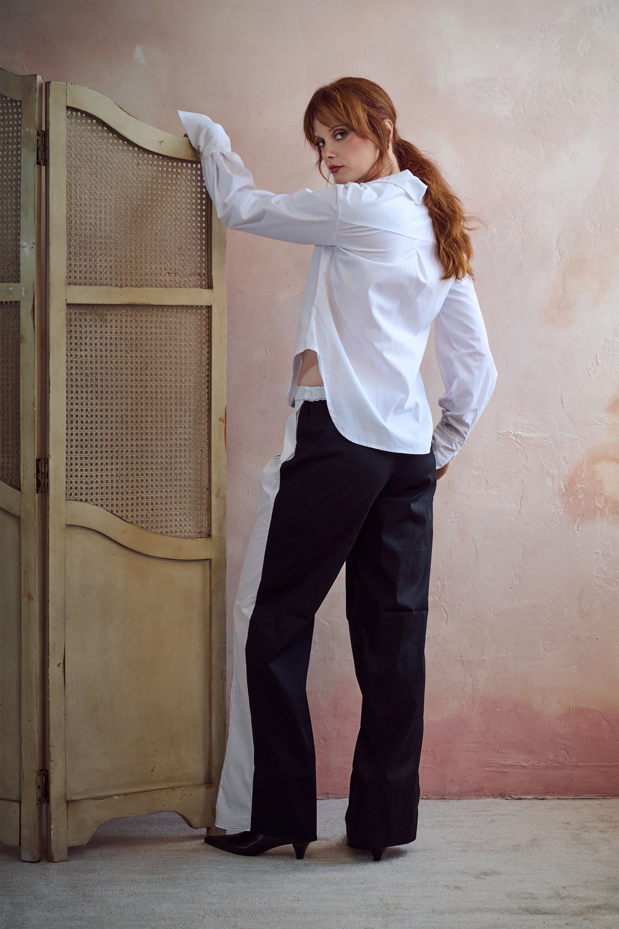 A woman with red hair stands in a studio setting wearing a white long-sleeve button-up shirt and black wide-leg trousers, leaning against a beige folding screen with a textured pink wall behind her.