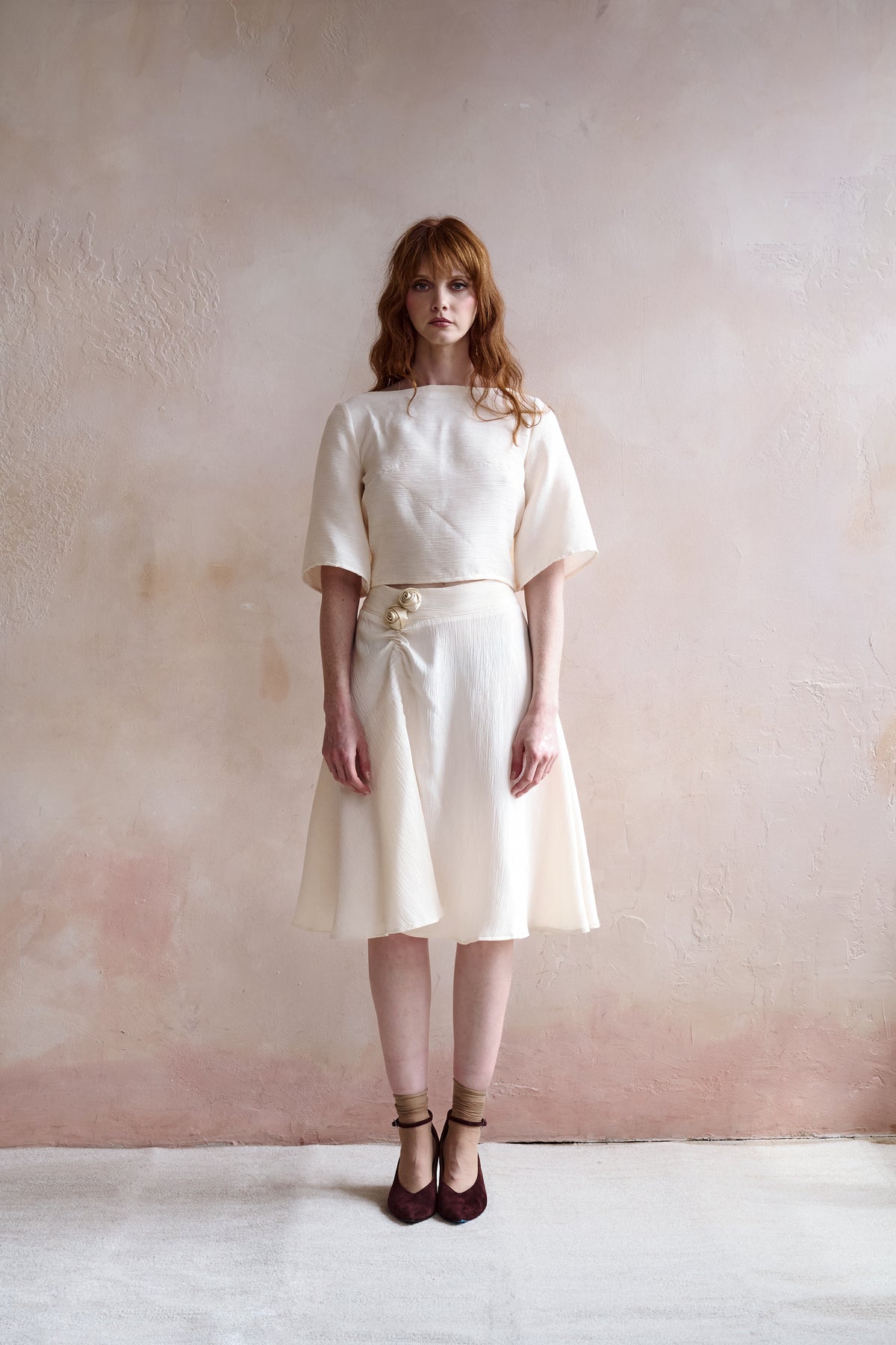 A woman with red hair stands against a textured, warm beige-pink wall wearing a cream-colored two-piece skirt suit with a short-sleeve top and matching pleated skirt, styled with a decorative belt and dark brown shoes.