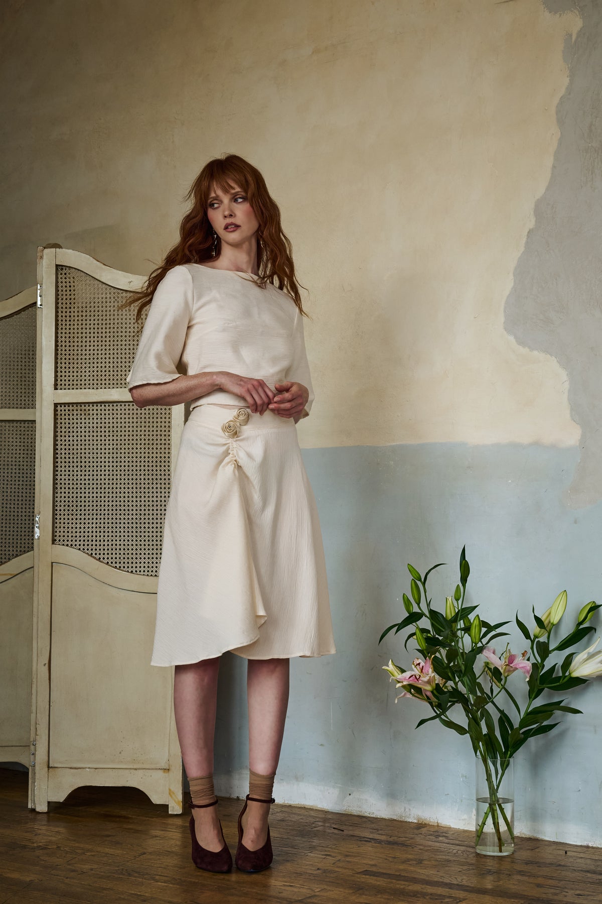 A woman stands in a studio setting wearing a cream-colored midi dress with a gathered waist and bow detail. The background features a textured plaster wall with a beige and blue color-blocked design, a wooden floor, a cane-screen partition, and a glass vase of flowers.