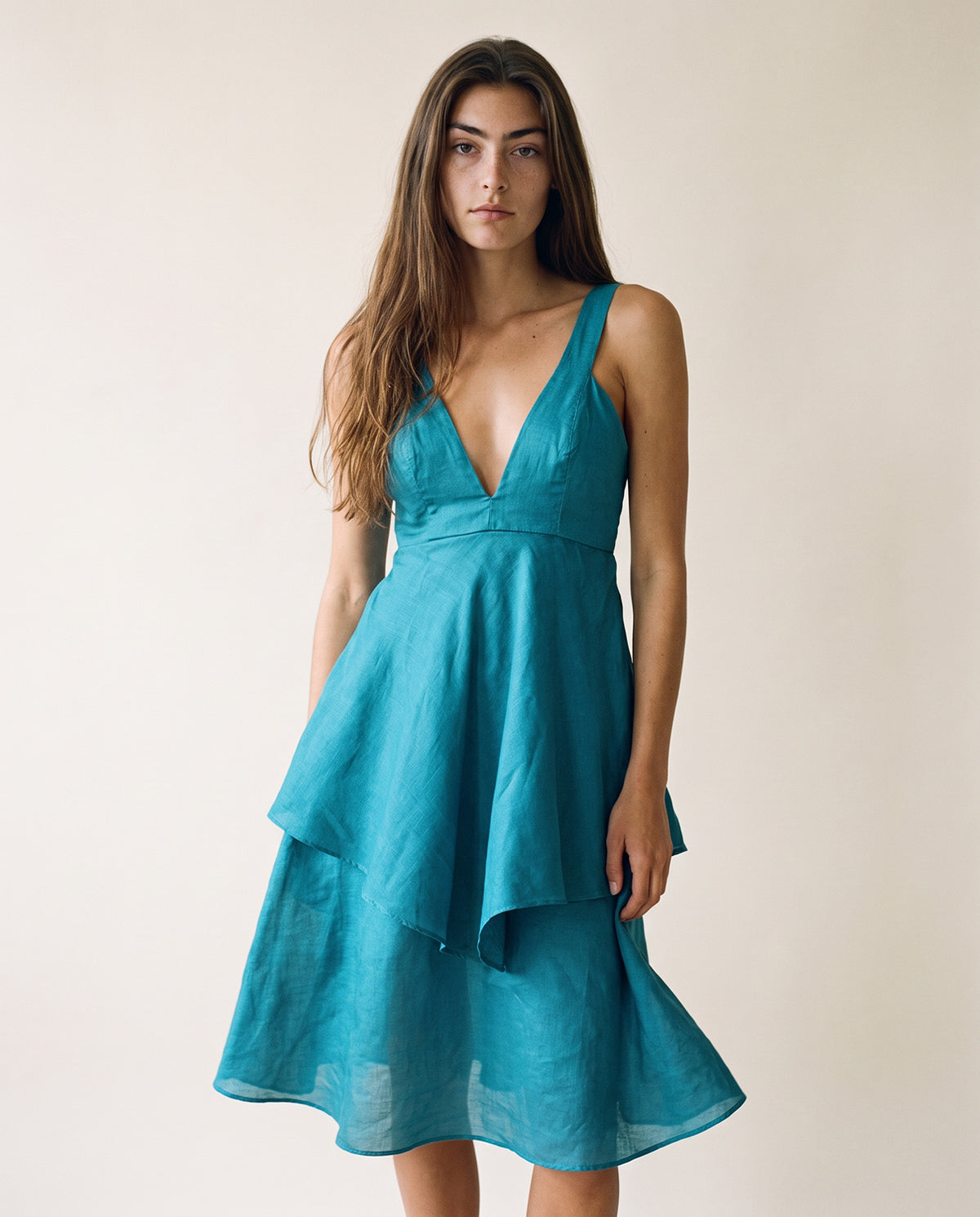 A woman modeling a teal blue-green sleeveless midi dress with a deep V-neckline and a layered, flared skirt against a plain beige background.