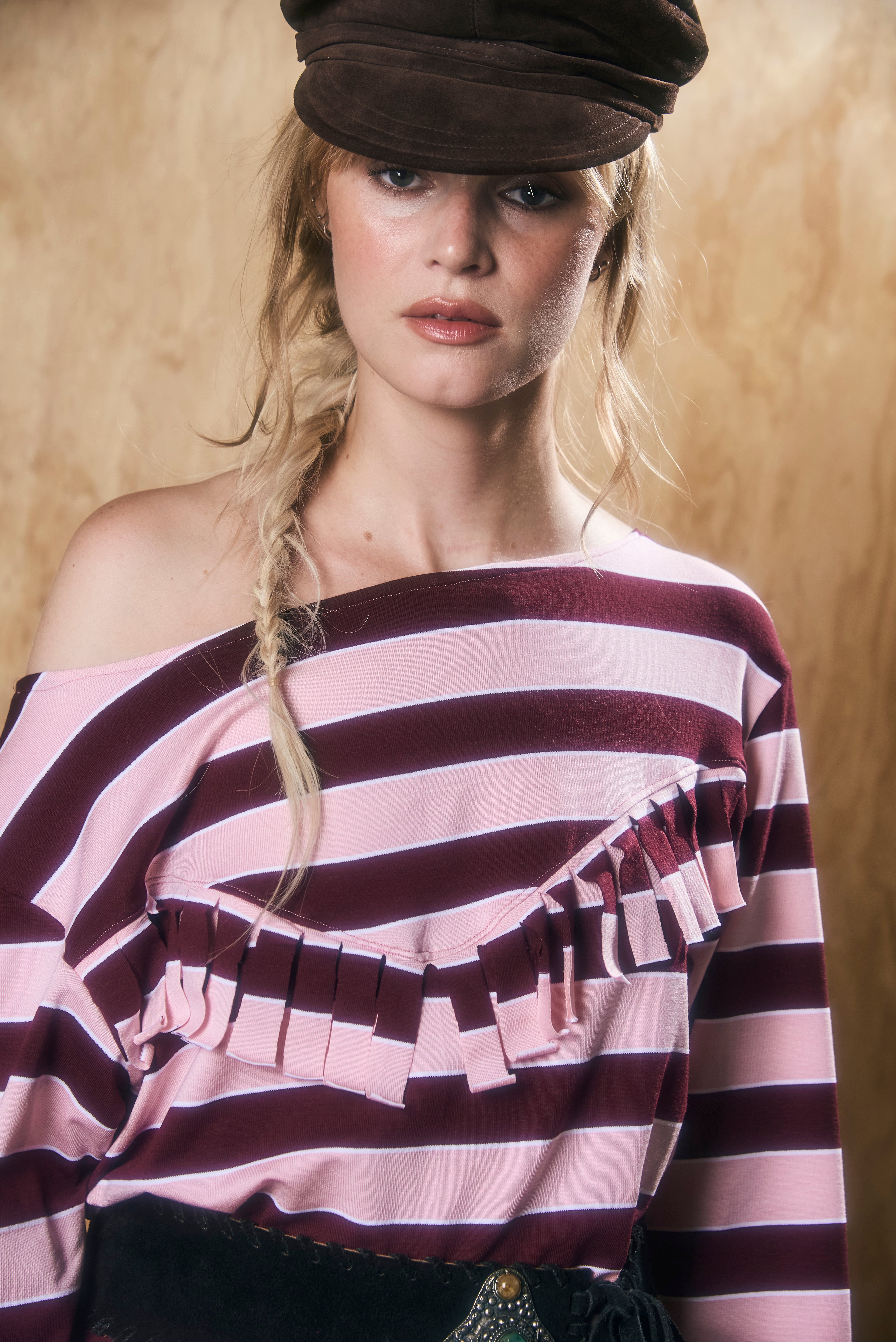 Woman wearing a striped one-shoulder dress with fringes and a cap against a wooden background