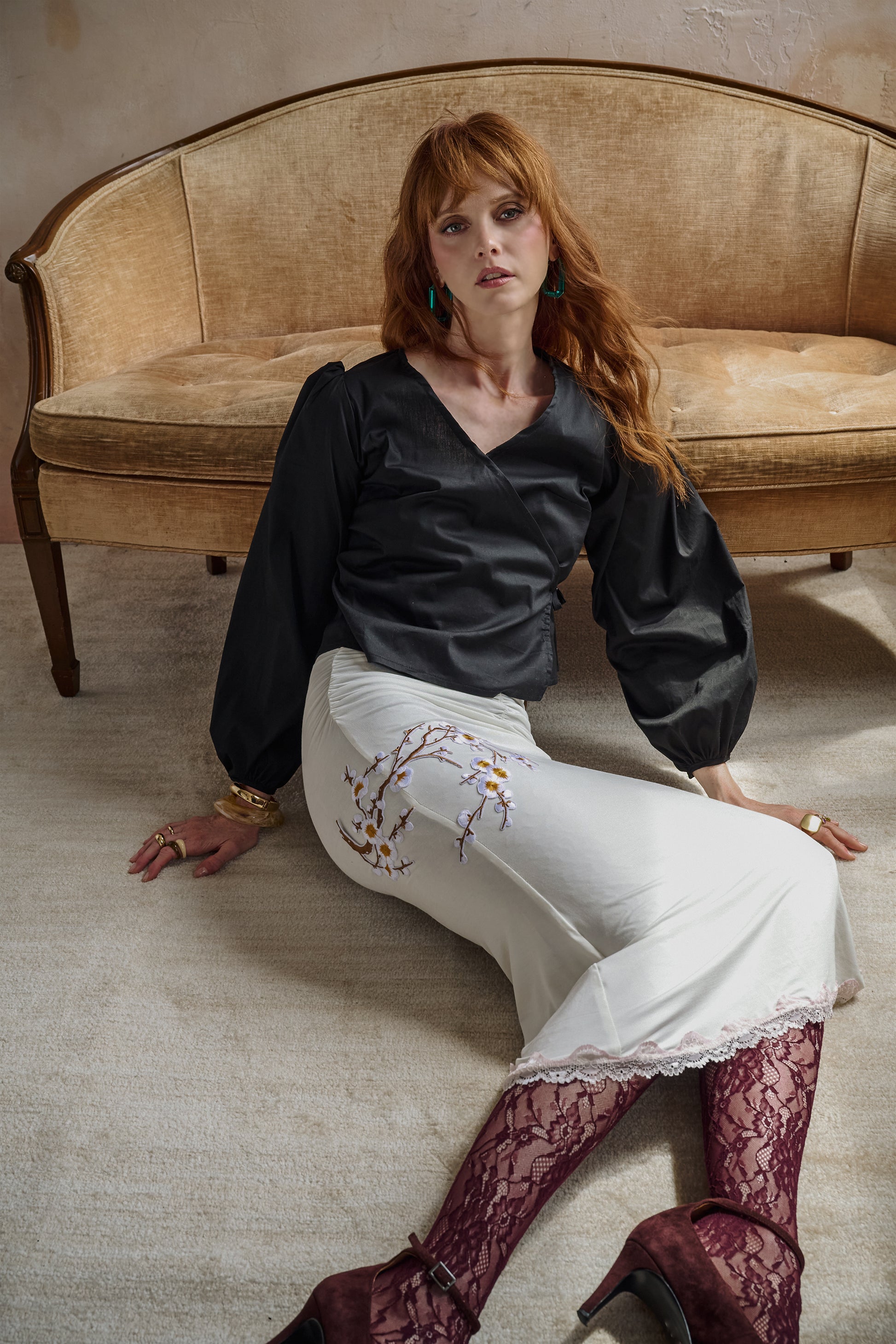 A woman with red hair sits on a beige carpet in front of a vintage-style sofa. She is wearing a black long-sleeve blouse, a white skirt with floral embroidery, burgundy lace tights, and dark red heels.