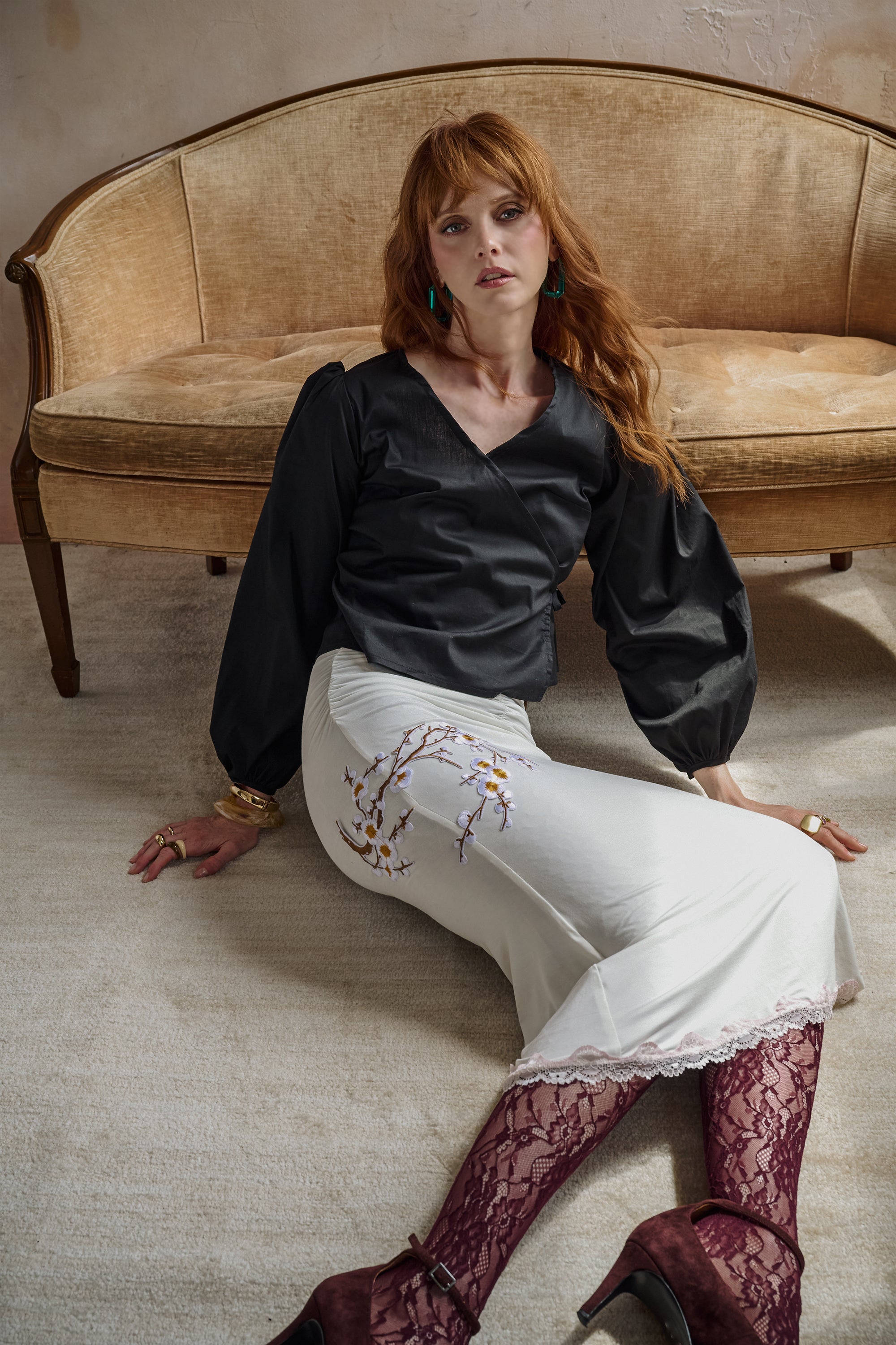 A woman with red hair sits on a beige carpet in front of a vintage-style sofa. She is wearing a black long-sleeve blouse, a white skirt with floral embroidery, burgundy lace tights, and dark red heels.