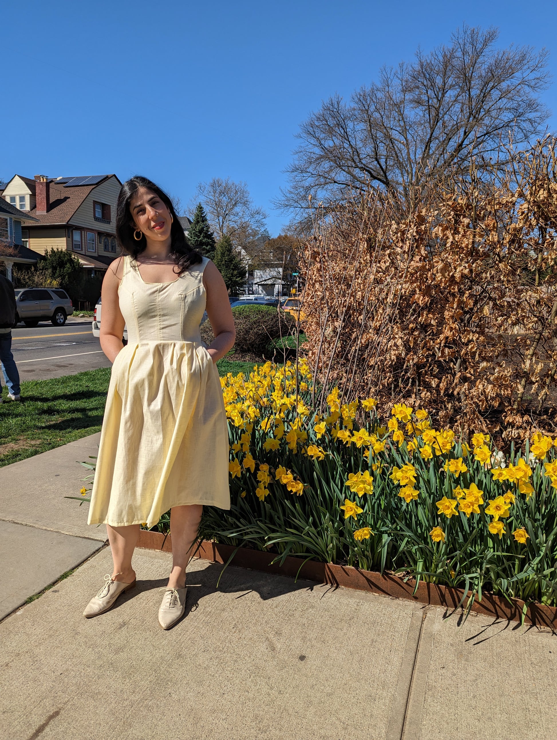 Woman in a yellow dress standing next to a flower bed with yellow flowers on a sidewalk.
