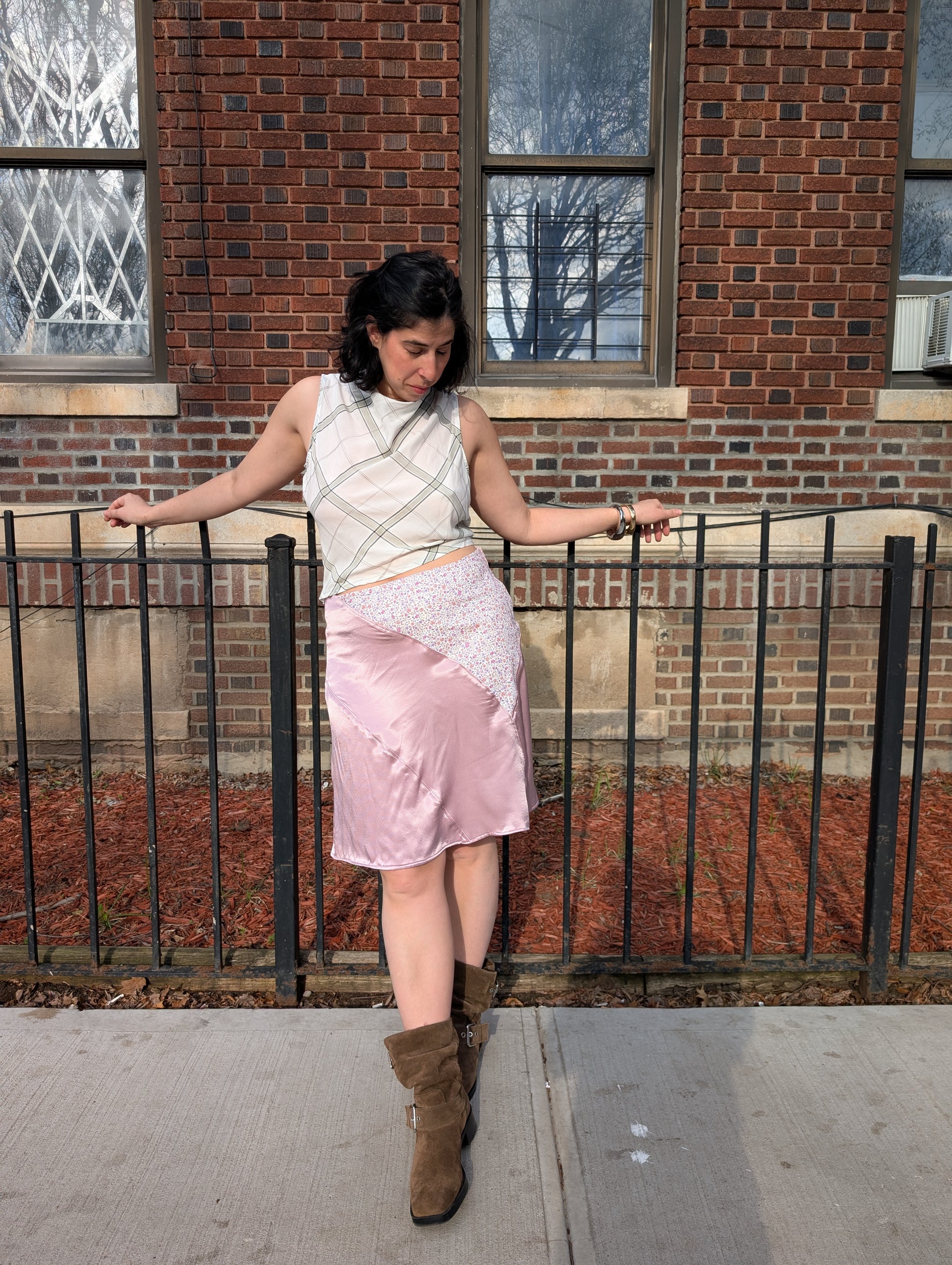Woman standing on a sidewalk wearing a white sleeveless top and pink skirt with brown boots, leaning against a railing.