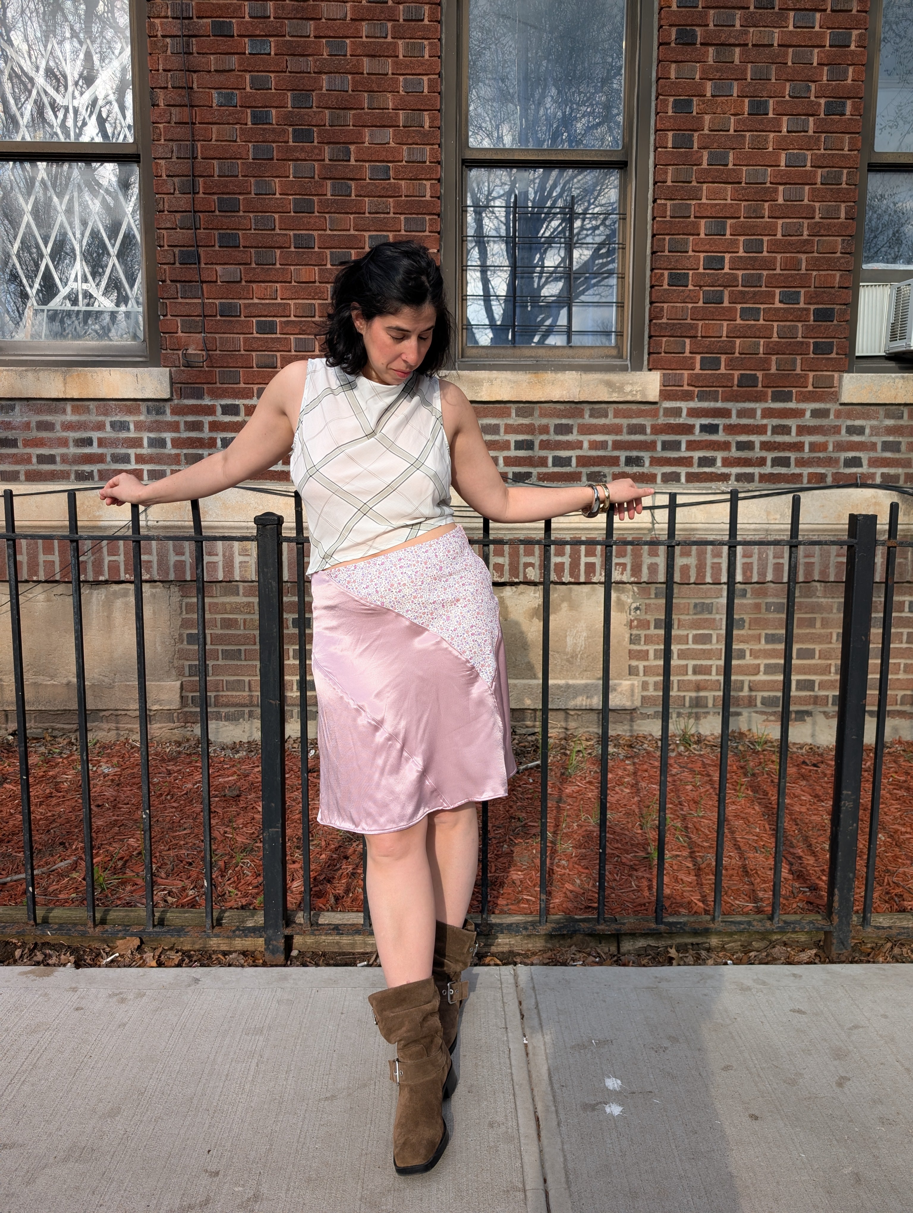 Woman standing on a sidewalk wearing a white sleeveless top and pink skirt with brown boots, leaning against a railing.