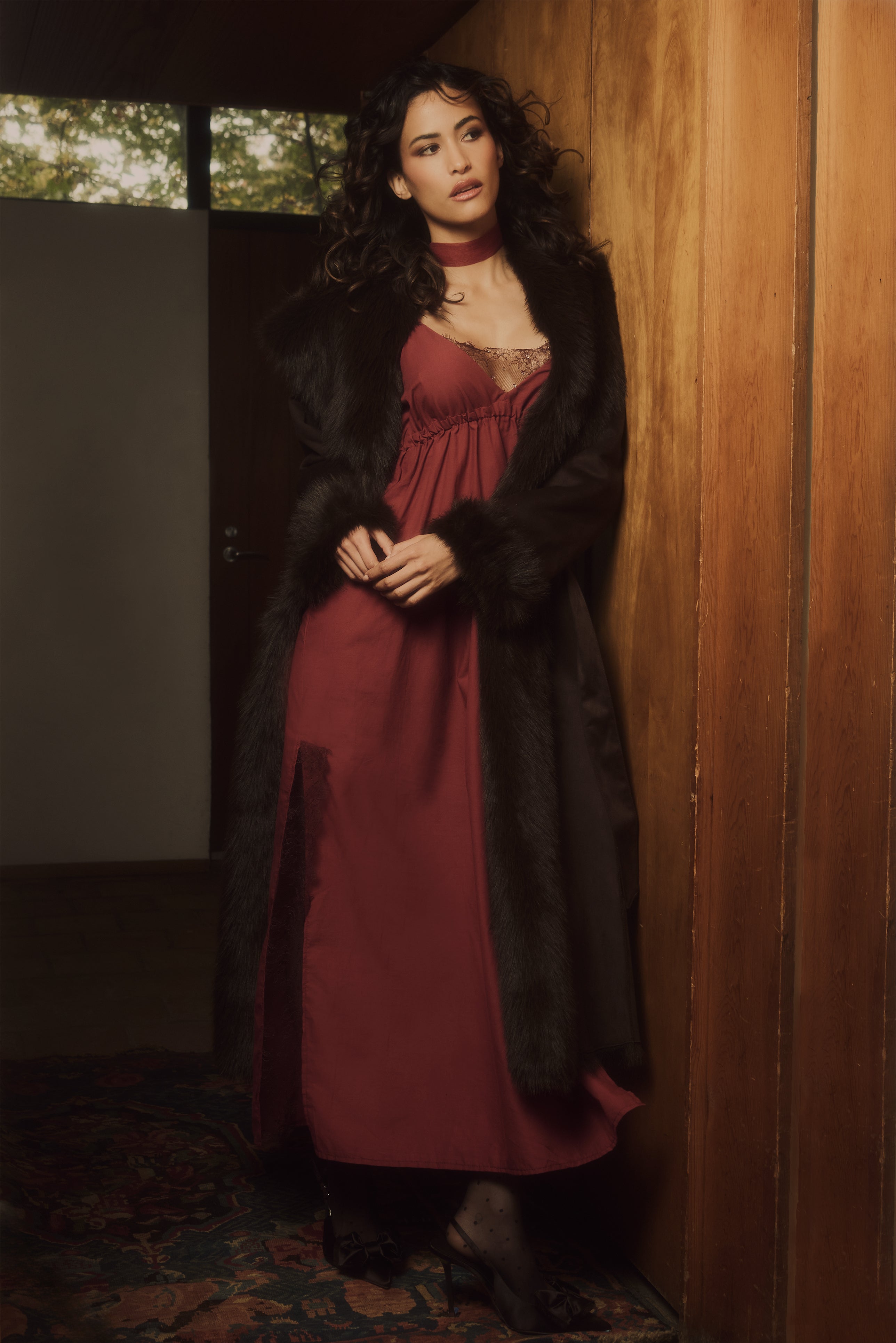 Woman in a long dark coat and red dress standing against a wooden wall.