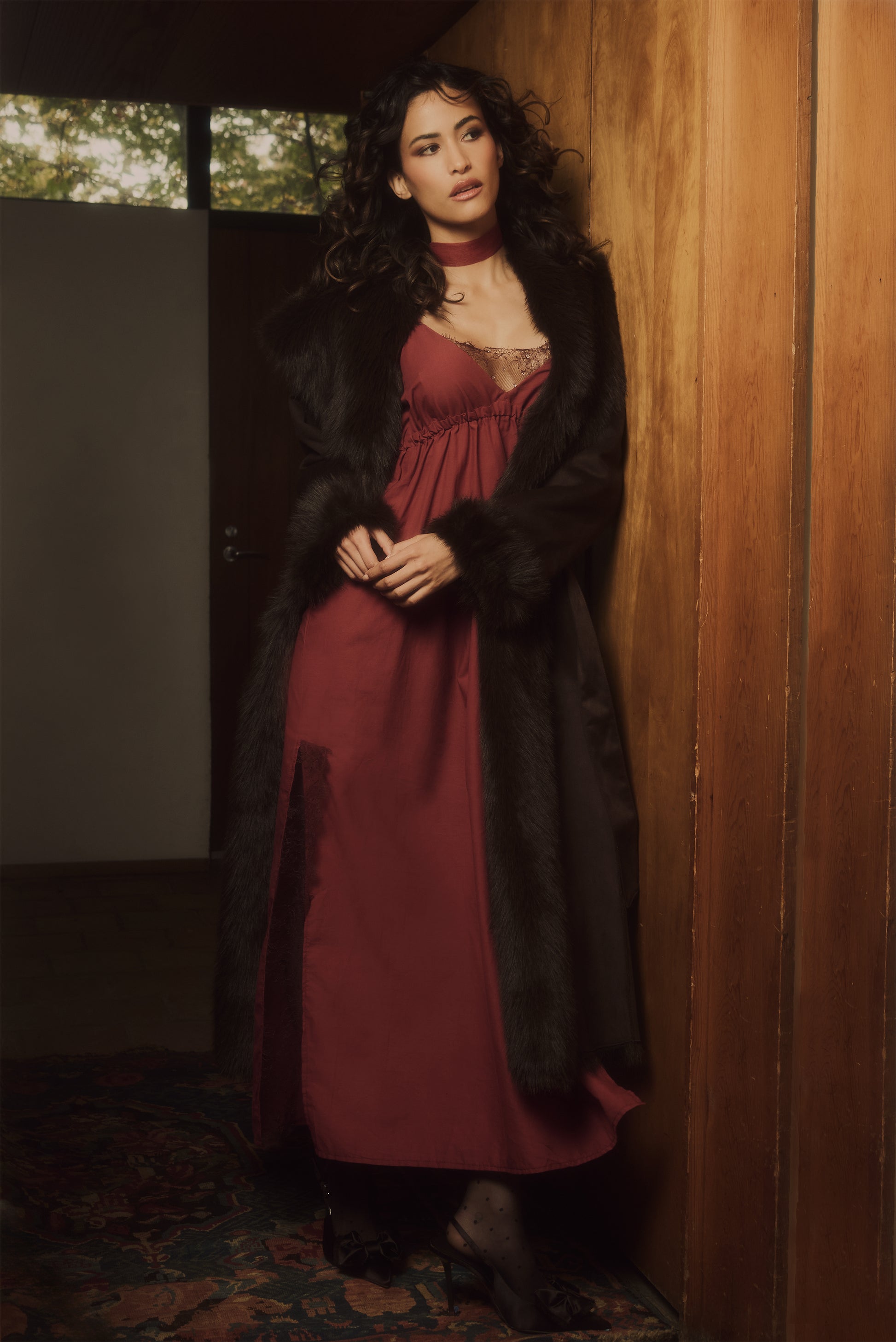 Woman in a long dark coat and red dress standing against a wooden wall.