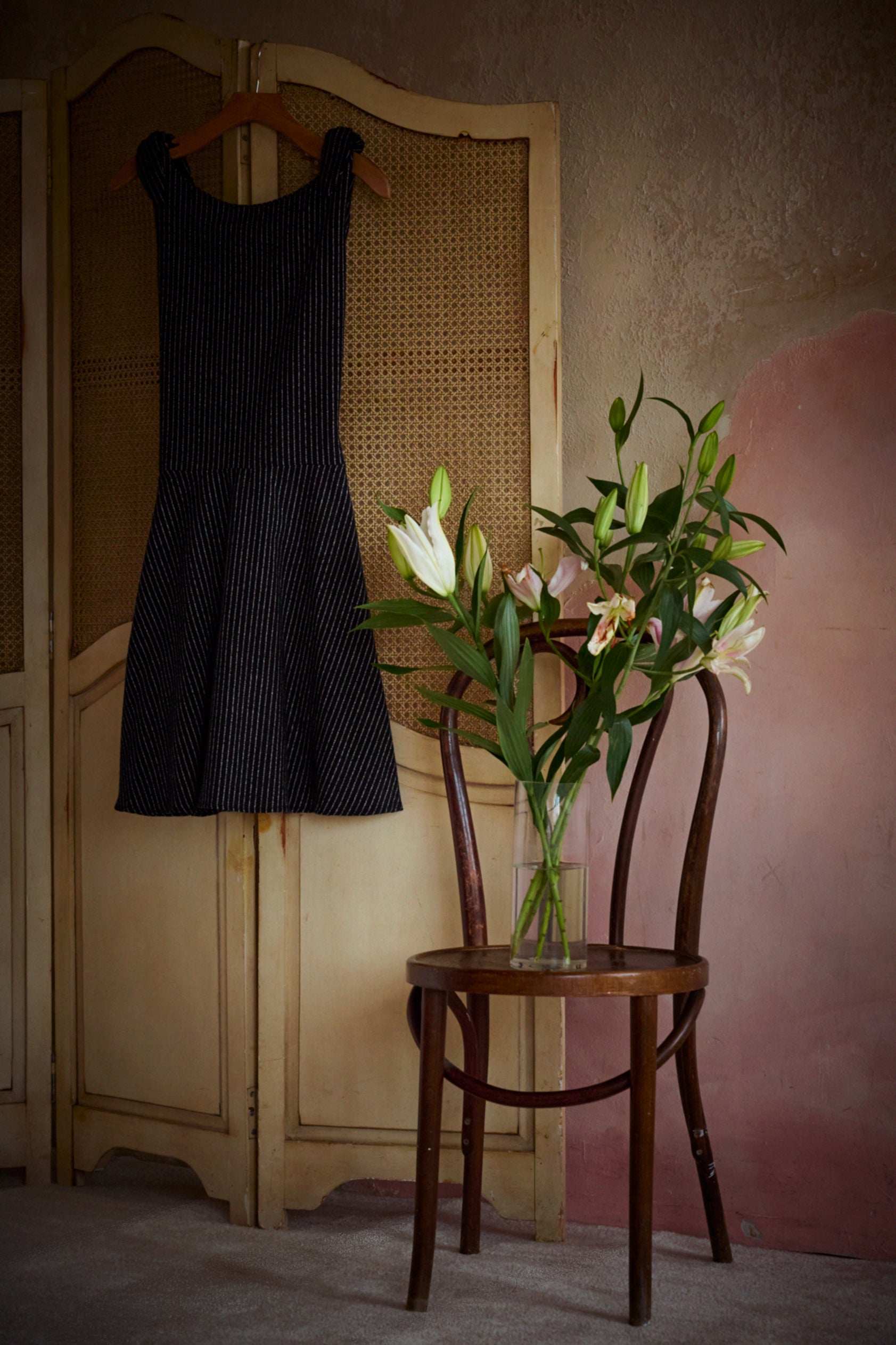 A sleeveless black pinstriped dress hangs on a wooden hanger against a beige cane-screen door, next to a wooden chair holding a vase of white lilies in a rustic room.