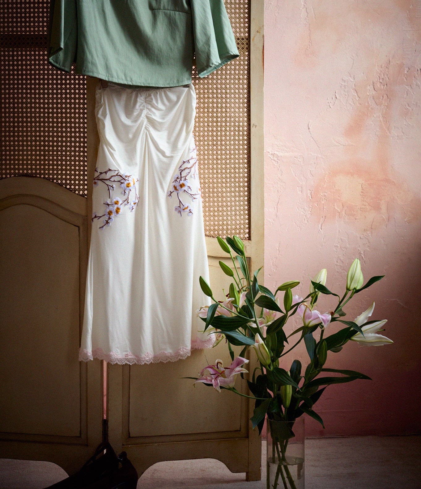 A sage green short-sleeve top and a white skirt with floral embroidery are displayed on a wooden hanger and stand against a rustic beige screen and pink wall, with a vase of lilies in the foreground.
