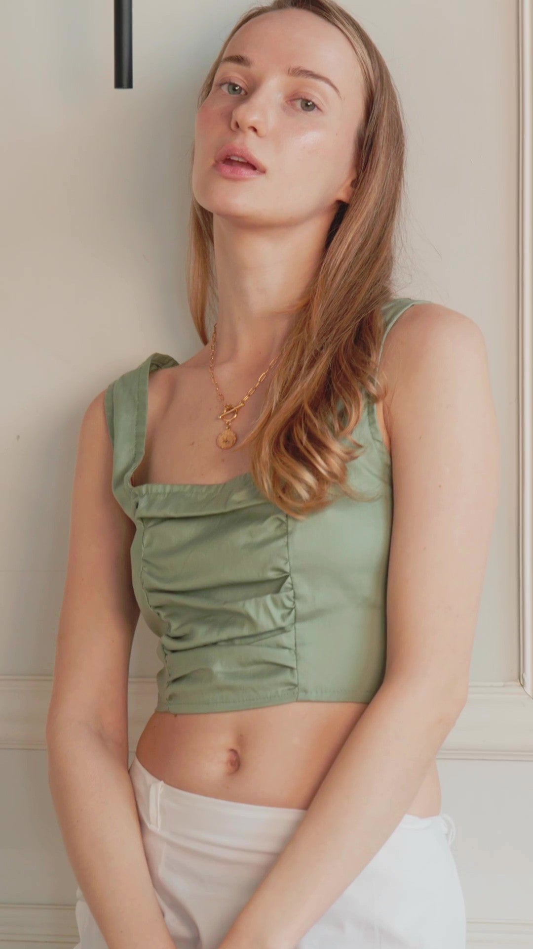 A woman wearing a sage green ruched crop top and white pants, posing against a light-colored paneled wall.