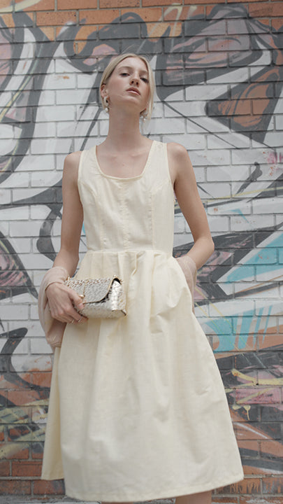 A woman wearing a sleeveless buttercream yellow colored cotton dress, holding a metallic clutch bag, standing in front of a graffiti-covered brick wall.