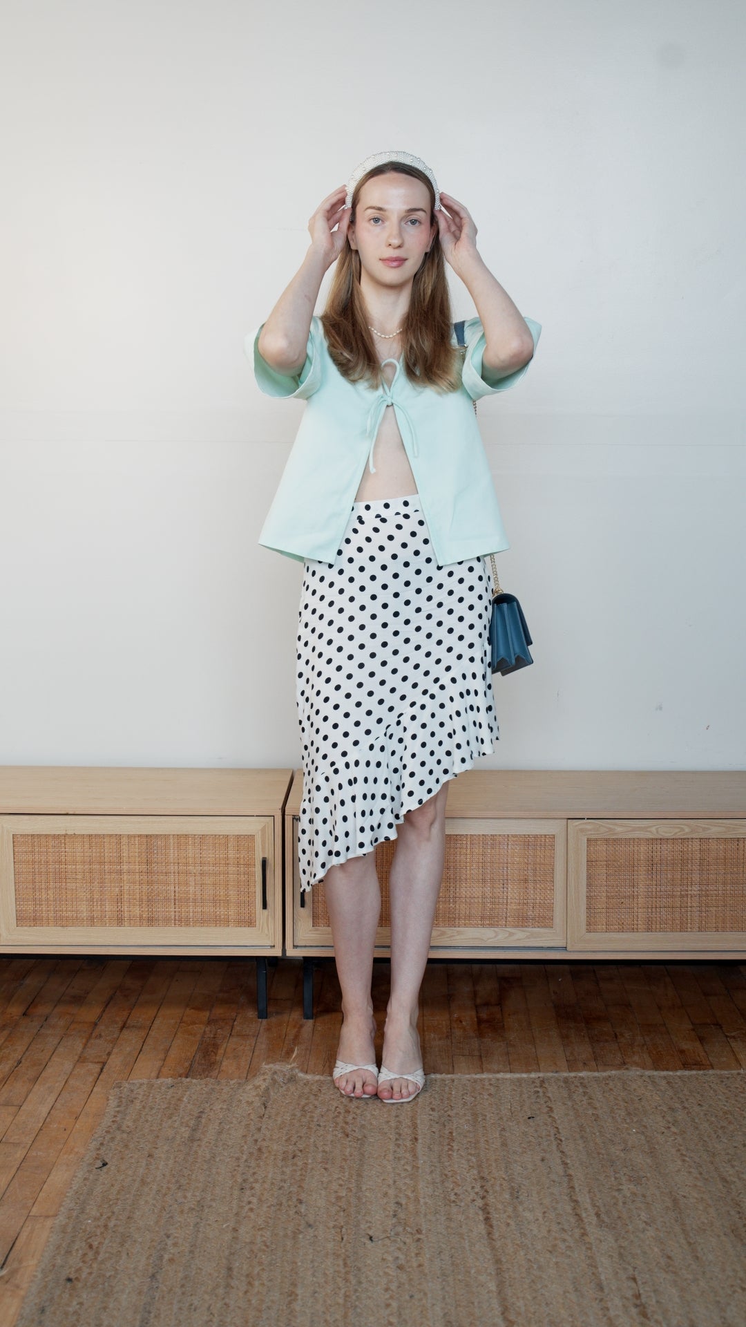 A woman modeling a mint green shirt and a black and white polka dot skirt, standing in a room with a light wood cabinet and wooden floor.