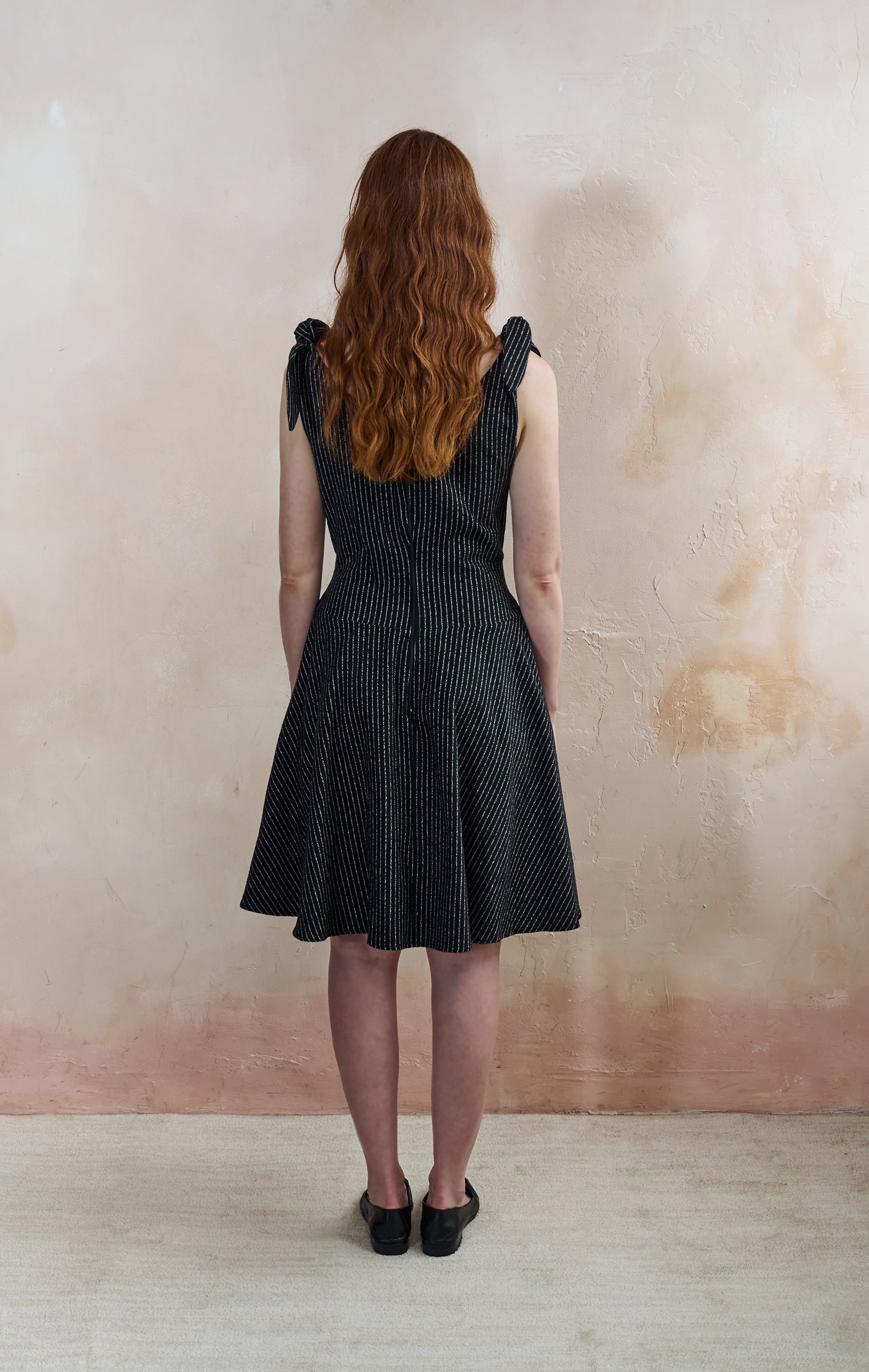A woman with long wavy red hair is shown from behind wearing a sleeveless black and white vertically striped dress with tie shoulder straps, standing against a textured beige plaster wall.