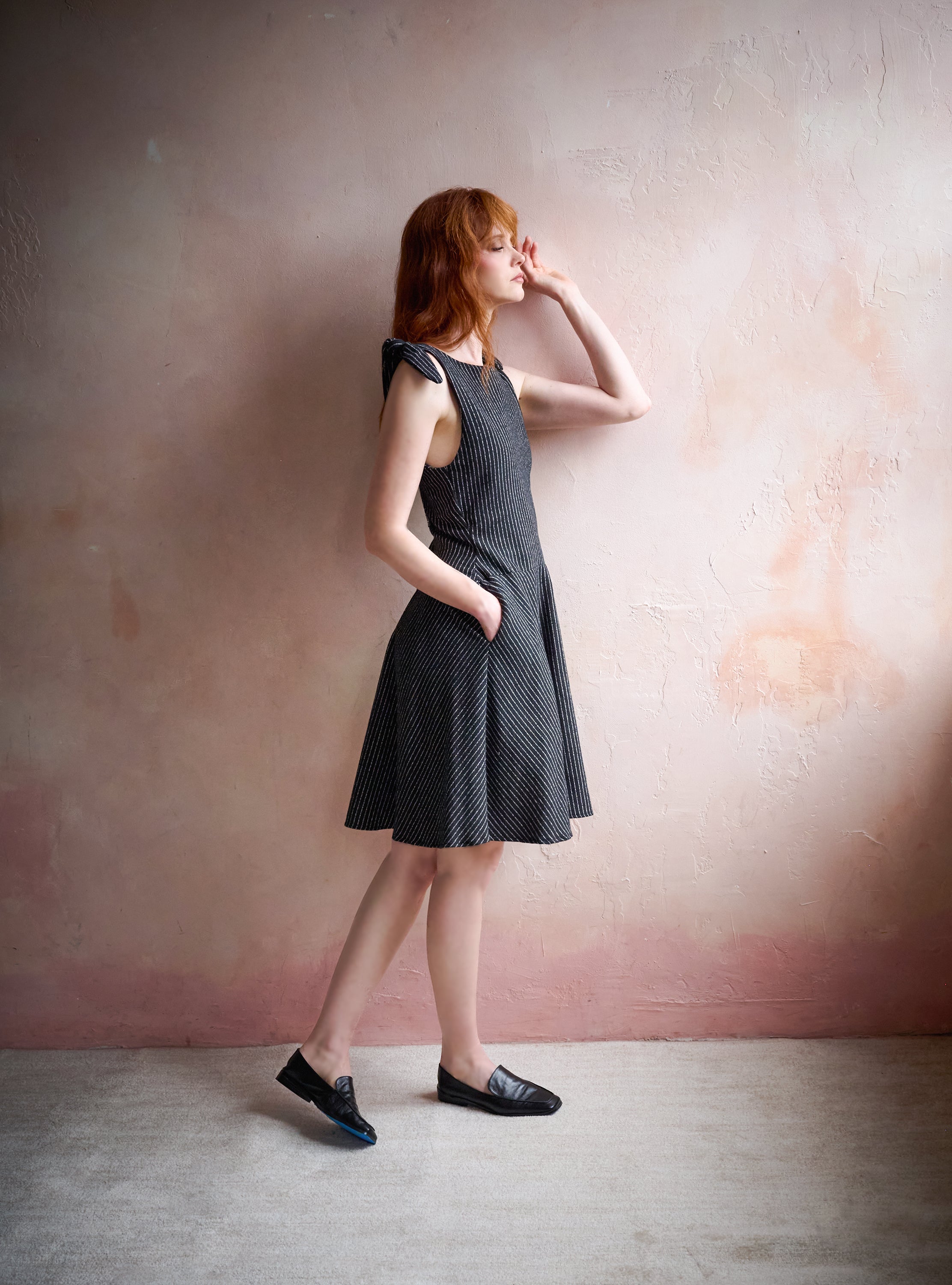 A woman with red hair models a sleeveless, knee-length black dress with a subtle white polka dot pattern, paired with black loafers, posing against a textured pinkish-beige wall.