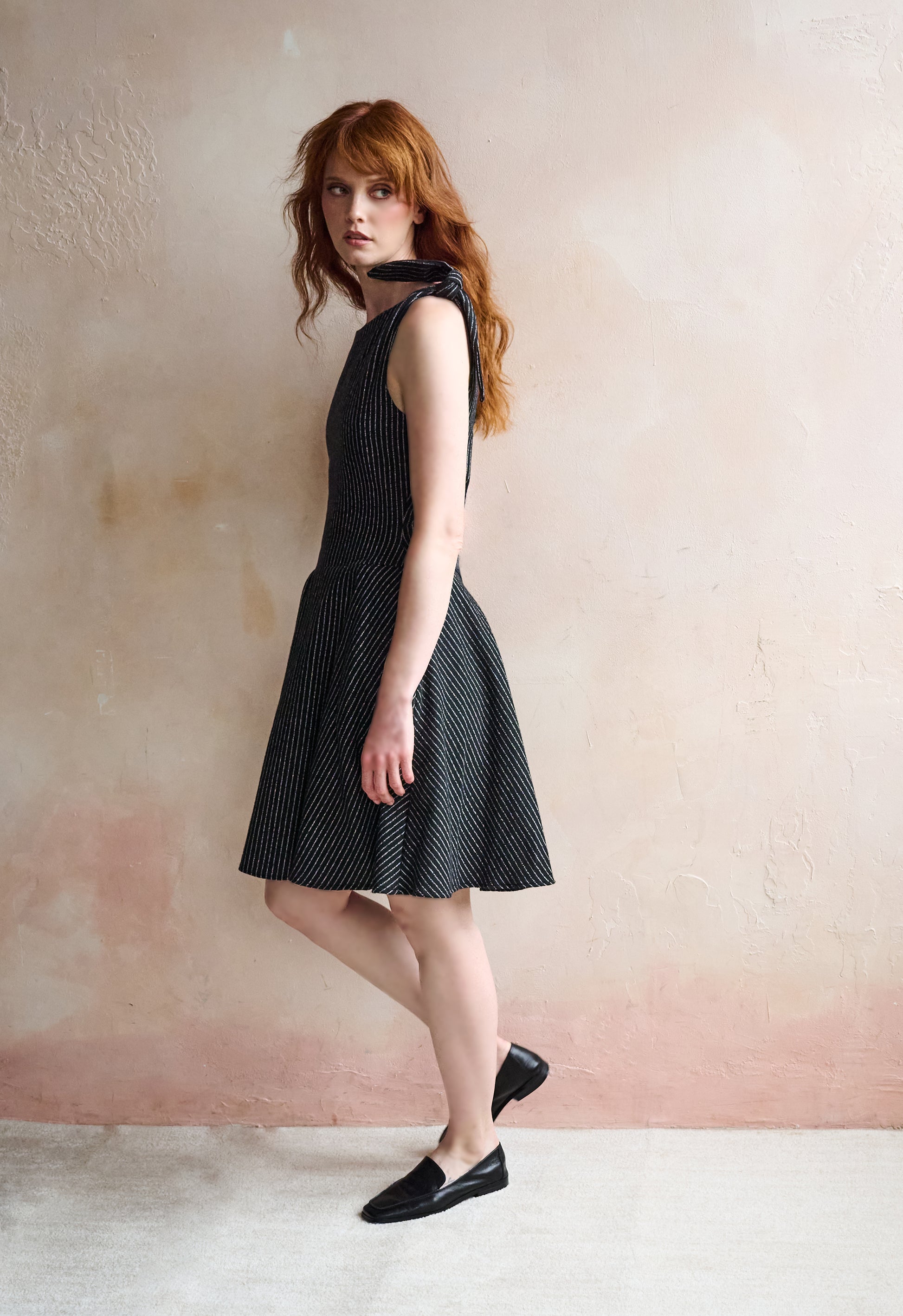 A woman with red hair models a sleeveless black and white micro-patterned dress with a tie-neck detail, paired with black loafers, standing against a textured pinkish-beige wall.