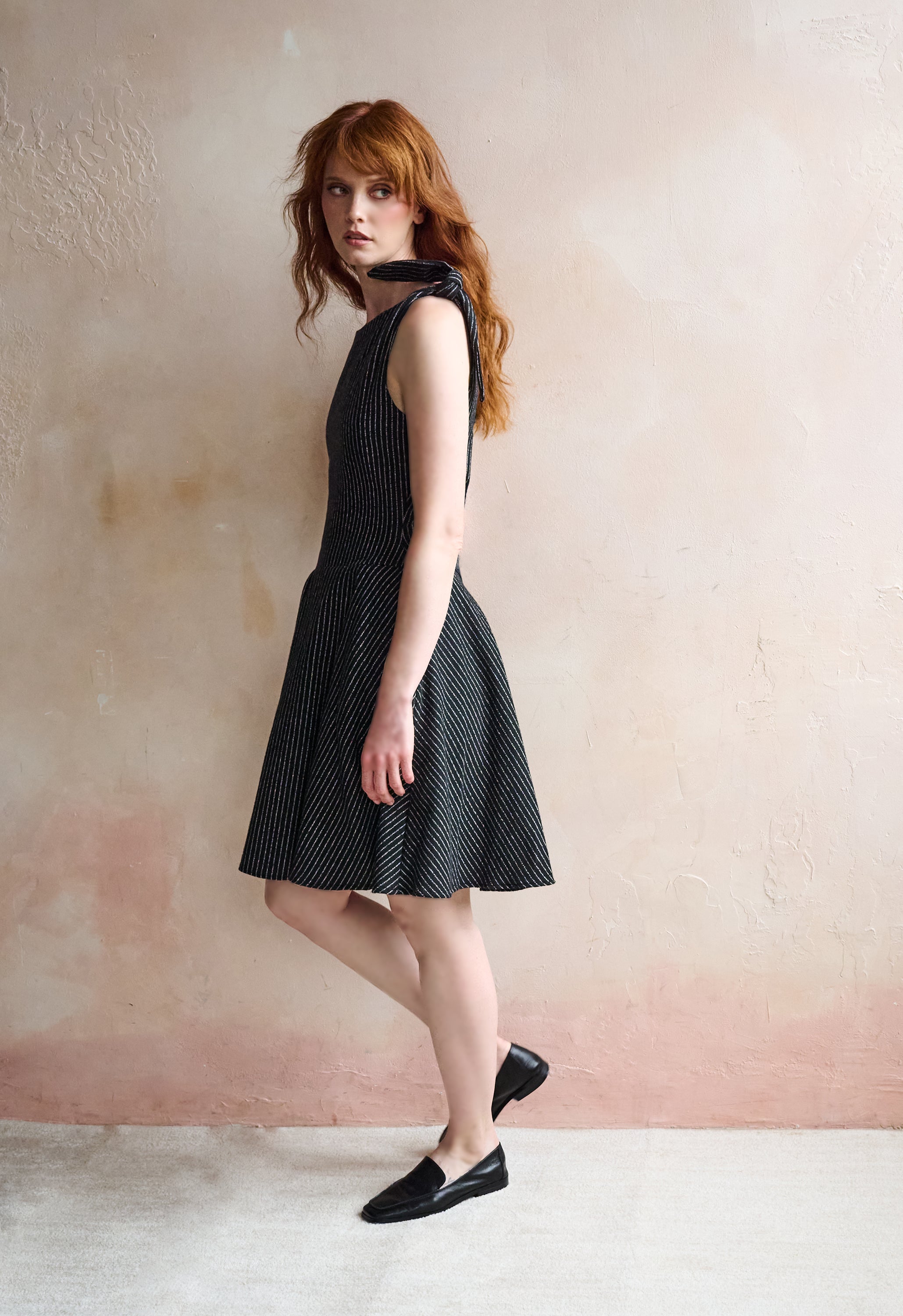 A woman with red hair models a sleeveless black and white micro-patterned dress with a tie-neck detail, paired with black loafers, standing against a textured pinkish-beige wall.
