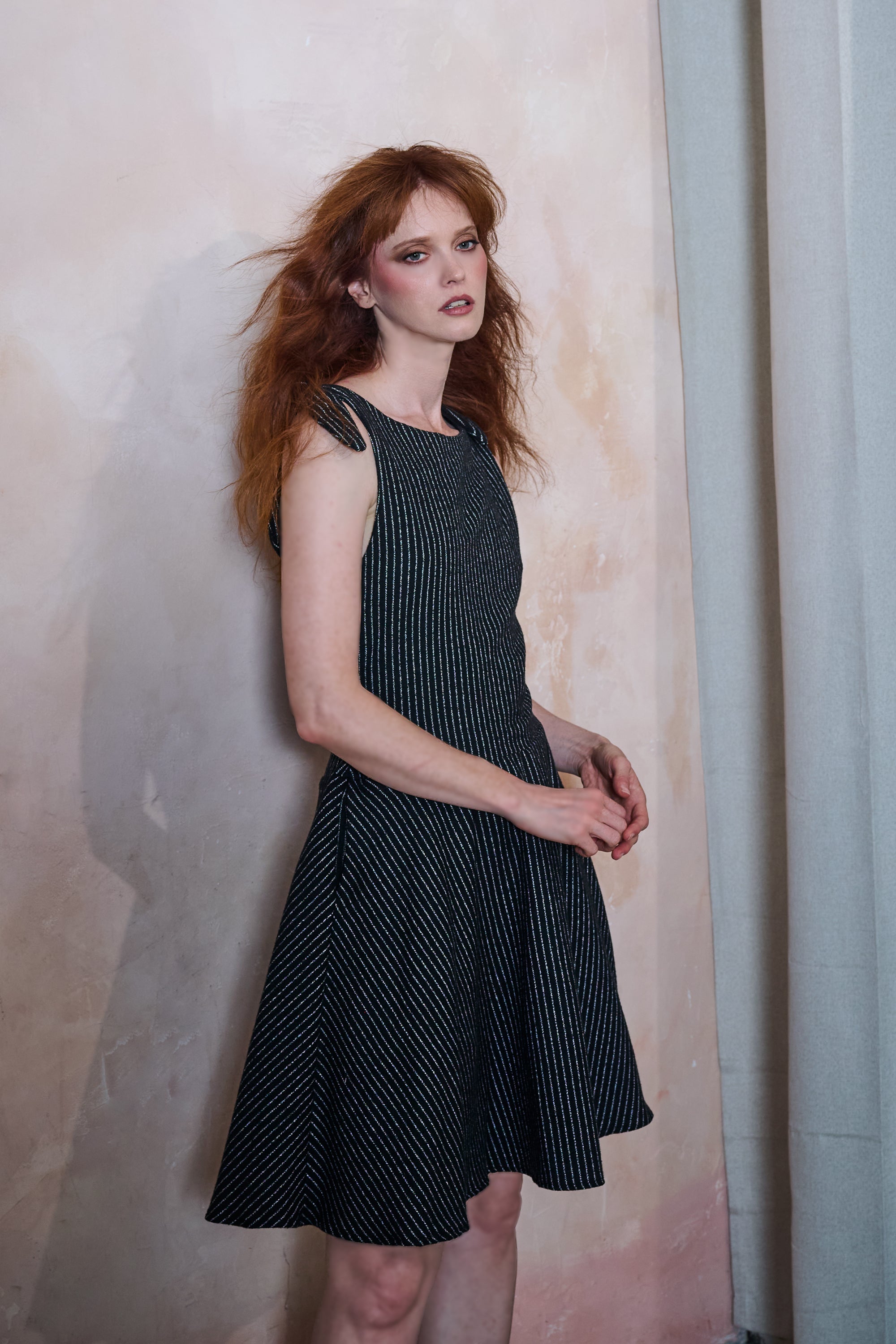 A woman with red hair wearing a sleeveless black dress with thin white vertical pinstripes, standing against a textured beige backdrop.